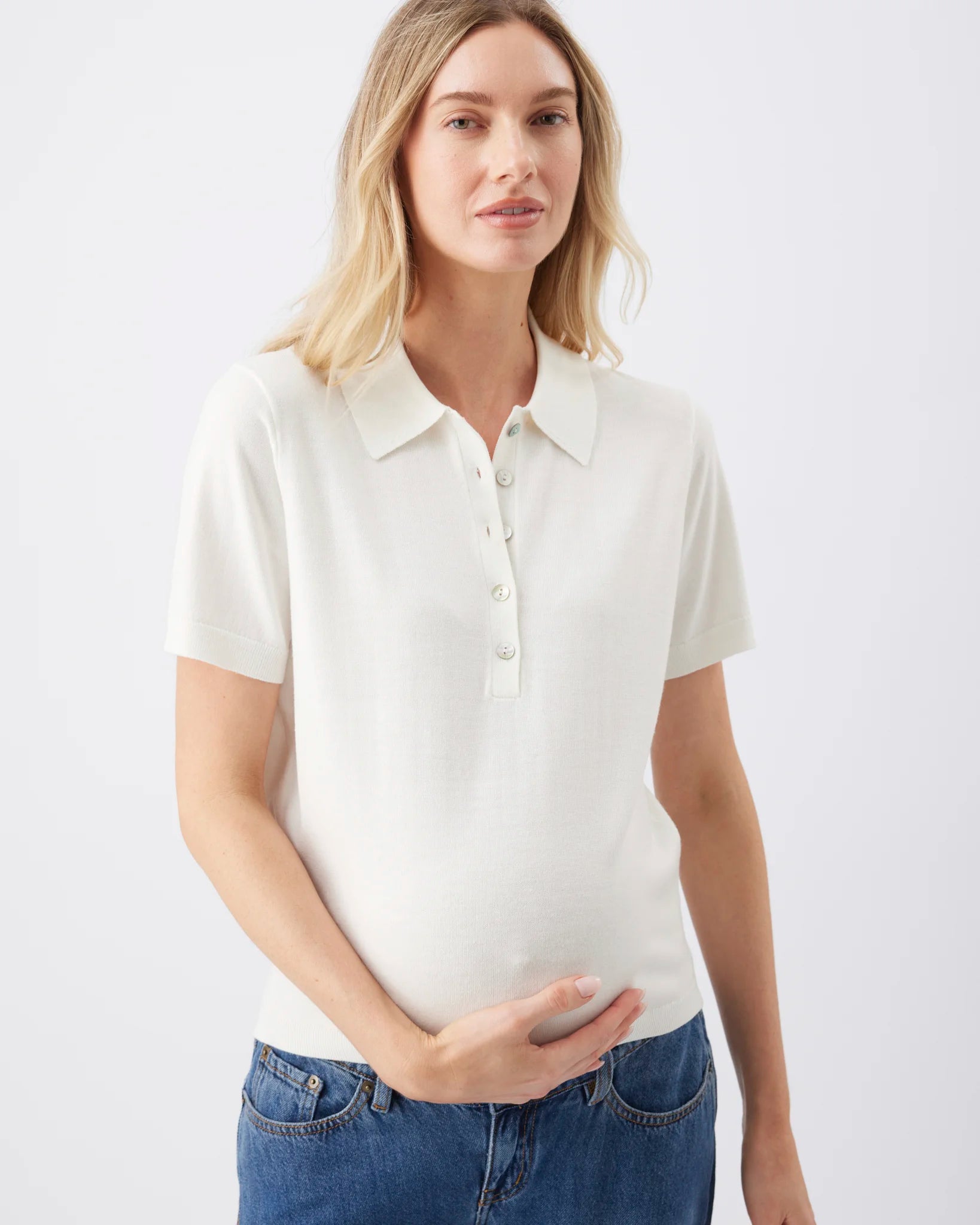 A woman wearing a short-sleeve polo shirt in off-white, paired with blue jeans, standing against a plain light gray studio background.