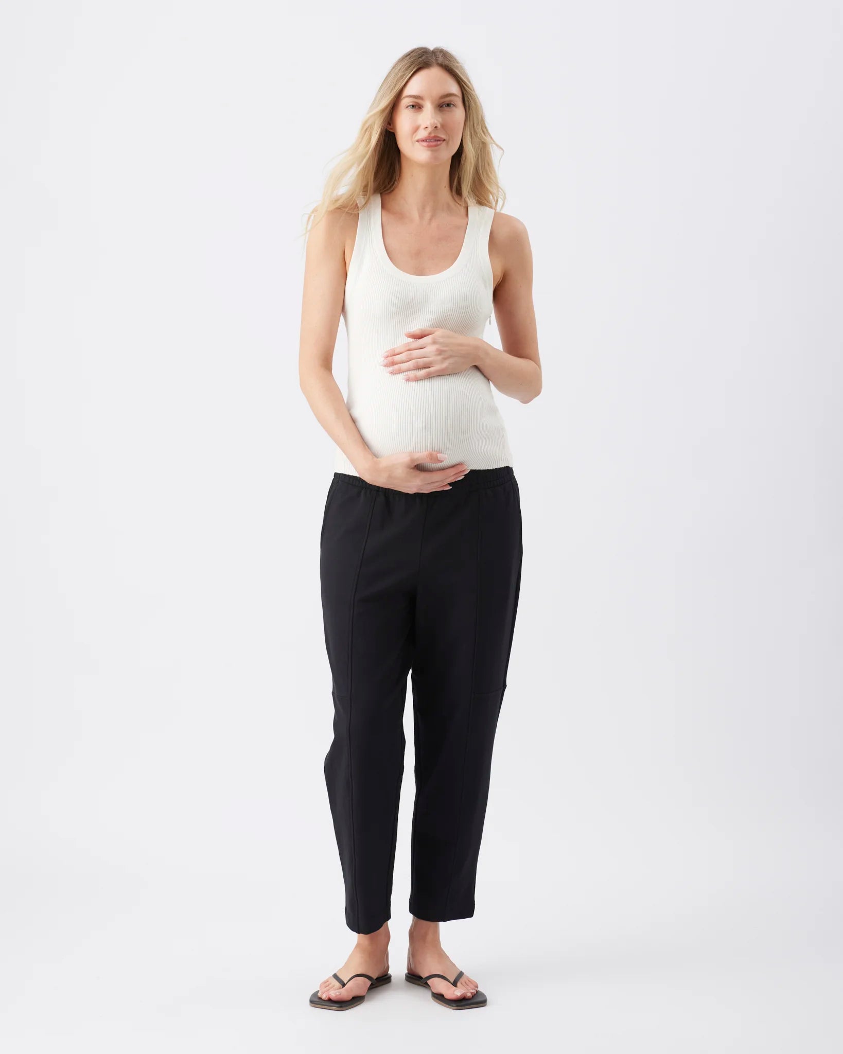 A pregnant woman stands against a light gray studio background wearing a white ribbed tank top, black cropped pants, and dark sandals, with her hands resting on her belly.