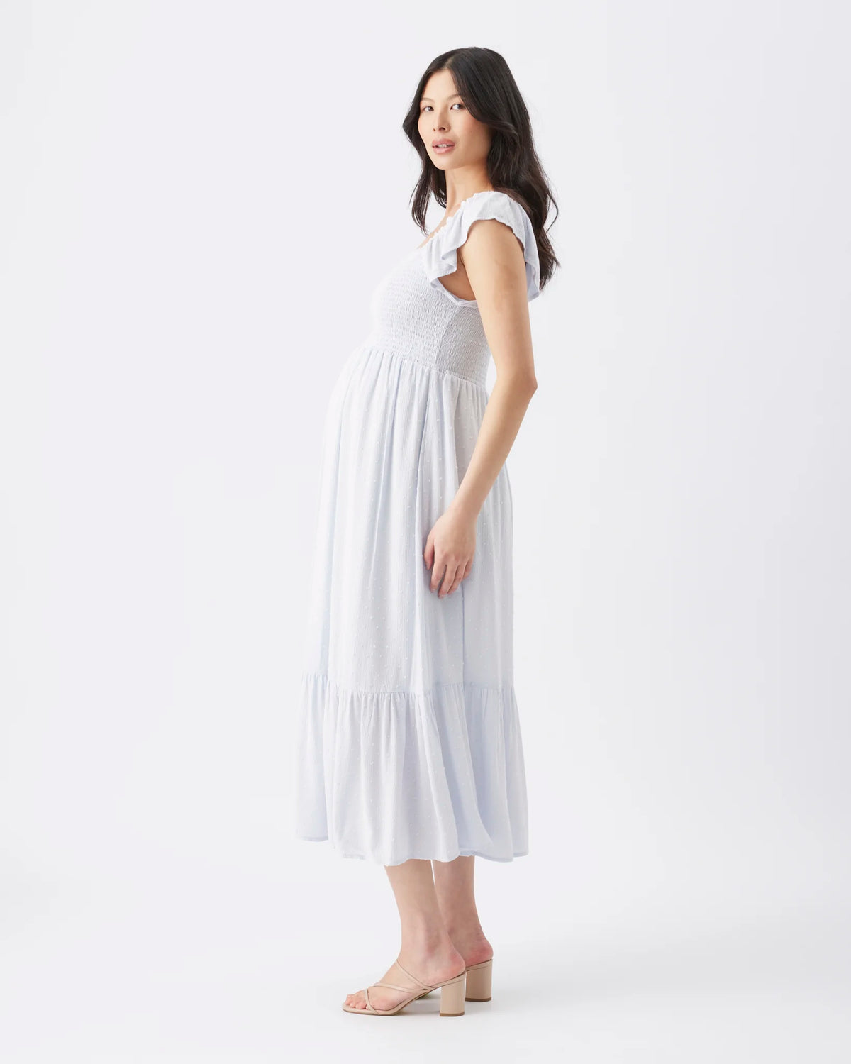 A pregnant woman wearing a white sleeveless midi maternity dress with ruffled shoulder straps and a tiered hem, standing in profile against a plain white studio background.