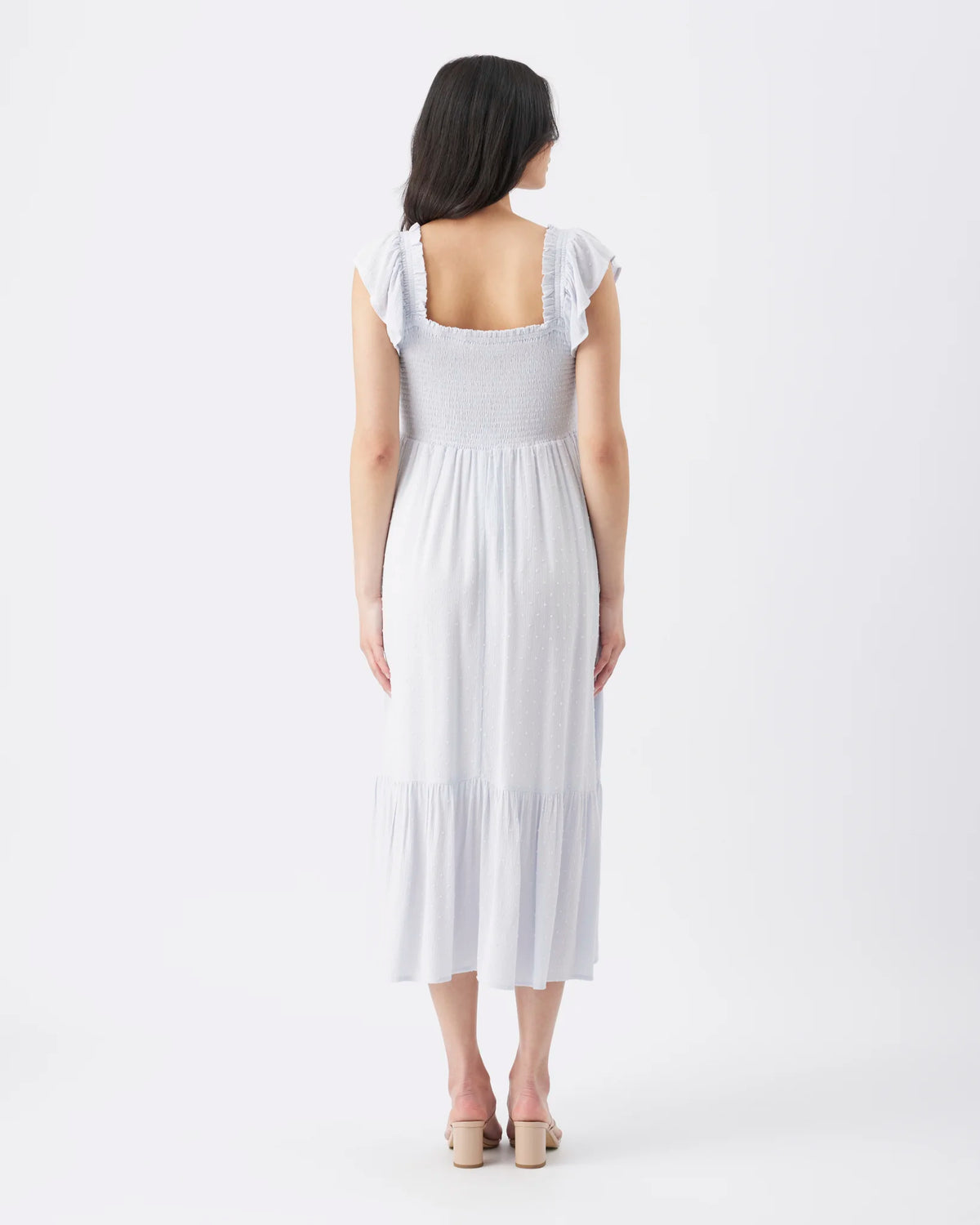 A woman viewed from the back wearing a white midi dress with ruffled cap sleeves, a square neckline, and a tiered skirt, paired with beige block-heel sandals against a plain white studio background.