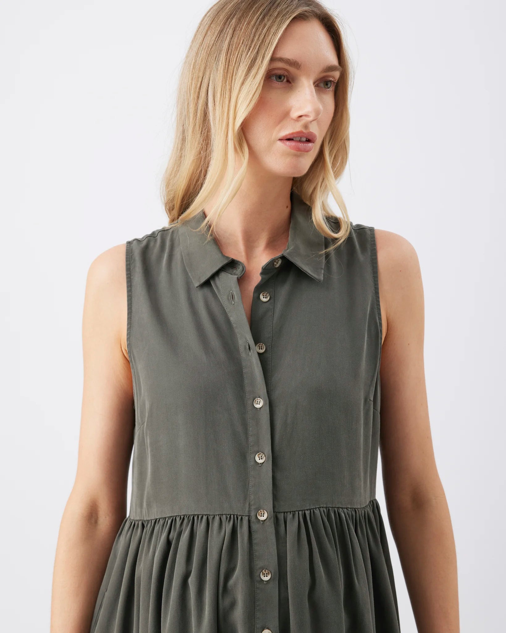 A woman wearing a sleeveless collared button-front dress in a muted olive green color, shown from the shoulders to the hips against a plain light gray studio background.