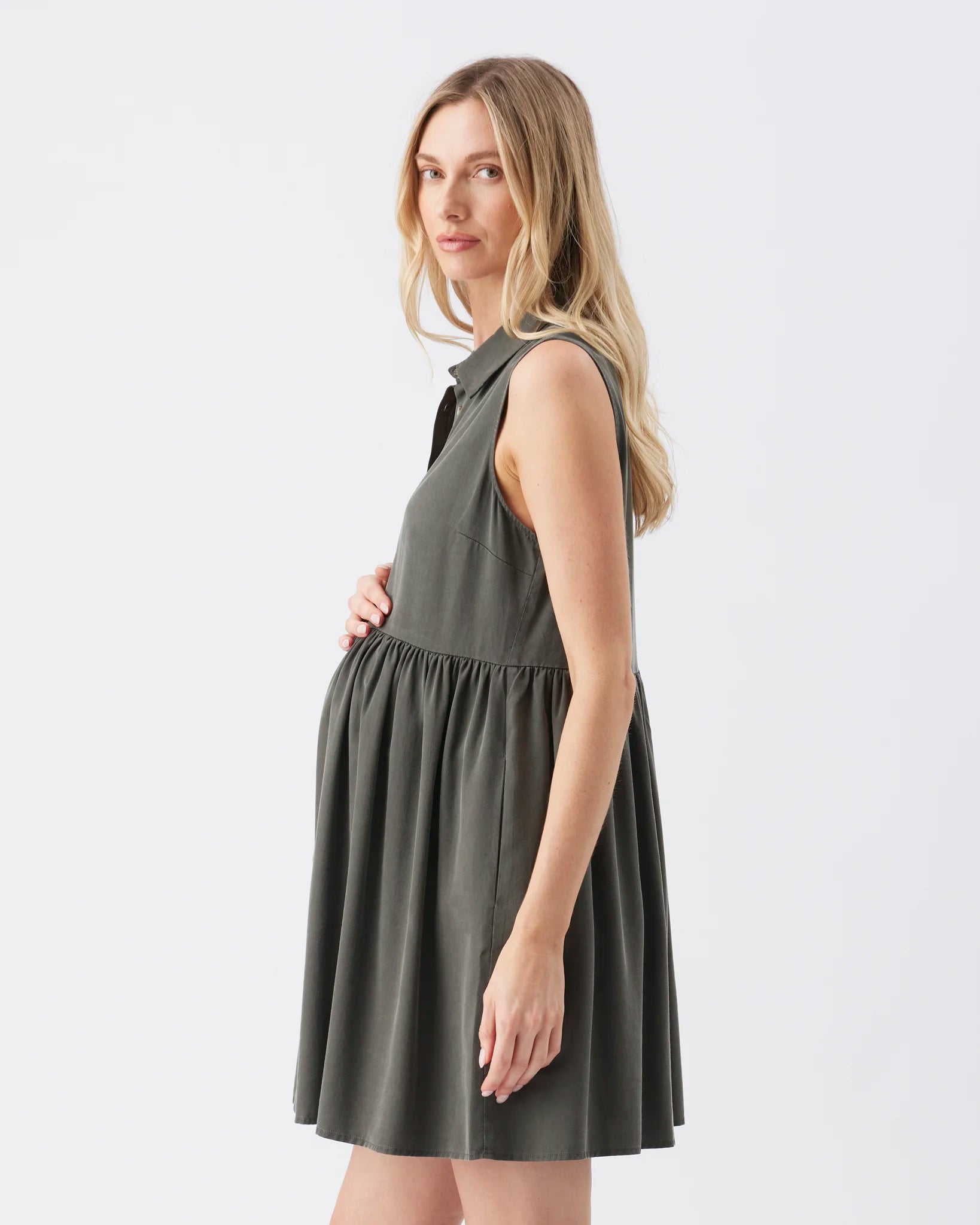 A pregnant woman wearing a sleeveless olive green maternity dress with a gathered waist, shown in profile against a plain white studio background.