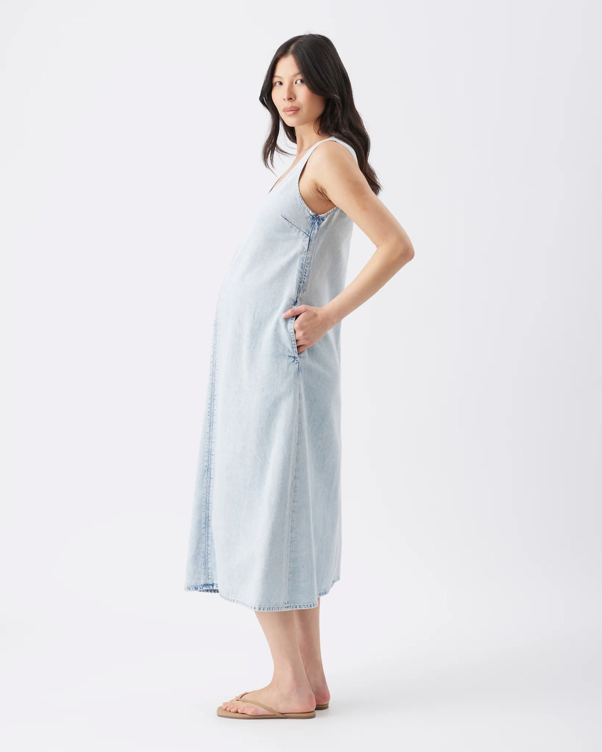 A pregnant model wearing a light blue denim maternity nightgown with side pockets, standing against a plain white studio background.