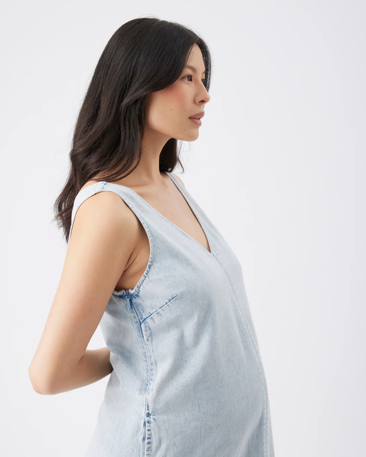 A woman in profile wearing a light blue denim sleeveless top against a plain white studio background.