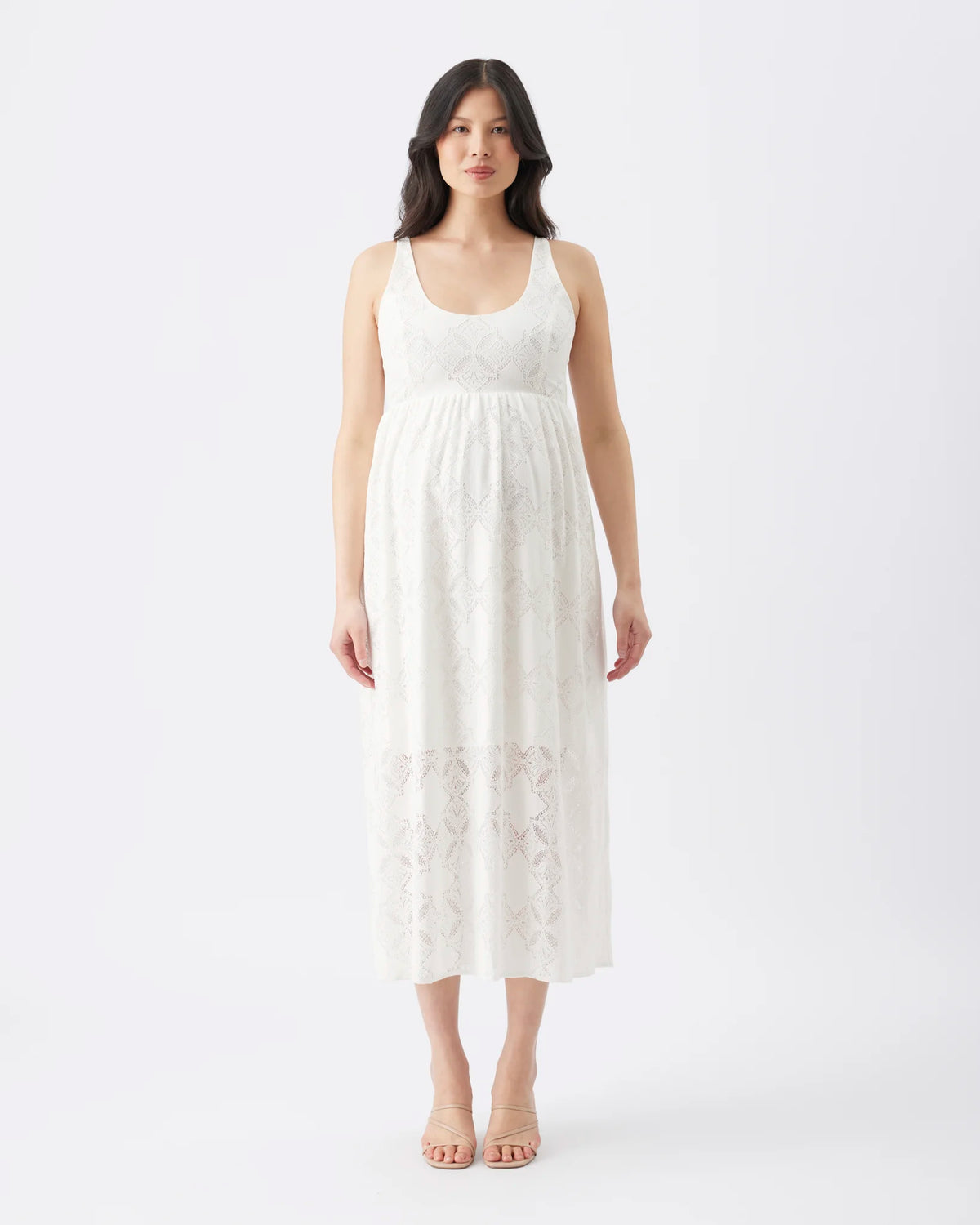 A woman modeling a sleeveless white midi dress with a scoop neckline and delicate lace pattern, standing against a plain white studio background.