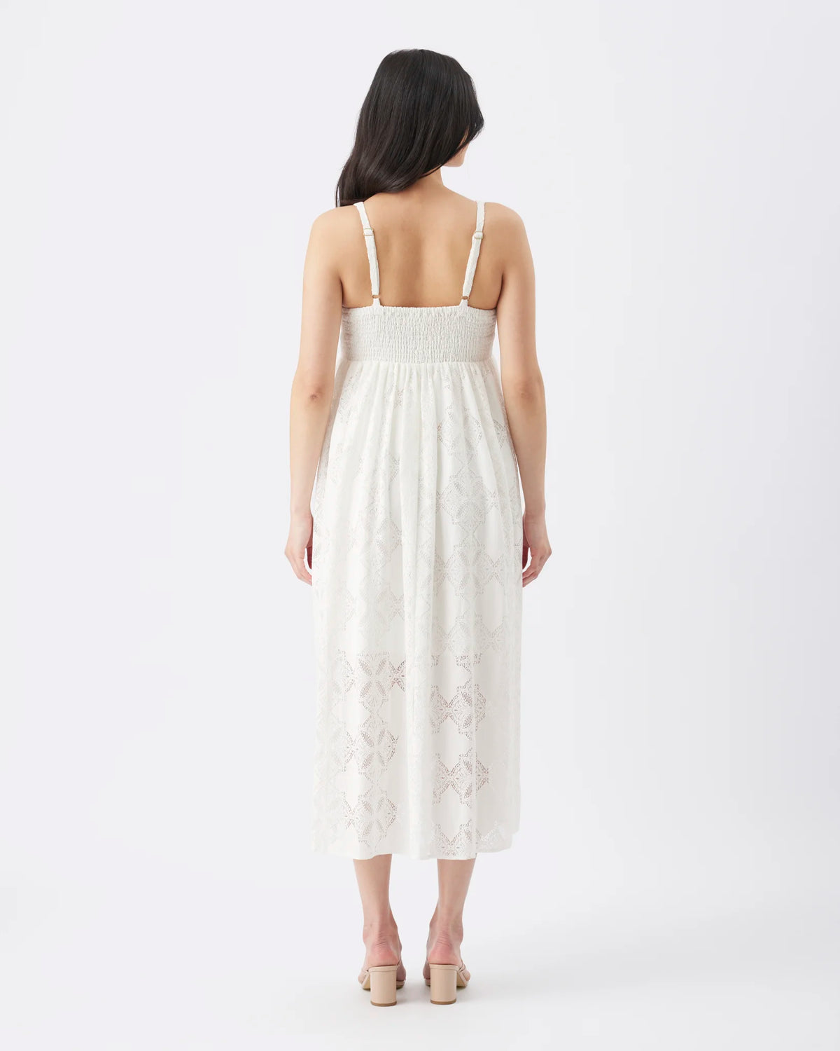 A woman viewed from the back wearing a white sleeveless midi dress with thin straps, a smocked waistband, and a subtle tonal floral lace pattern near the hem, paired with beige block-heel sandals on a light gray studio background.