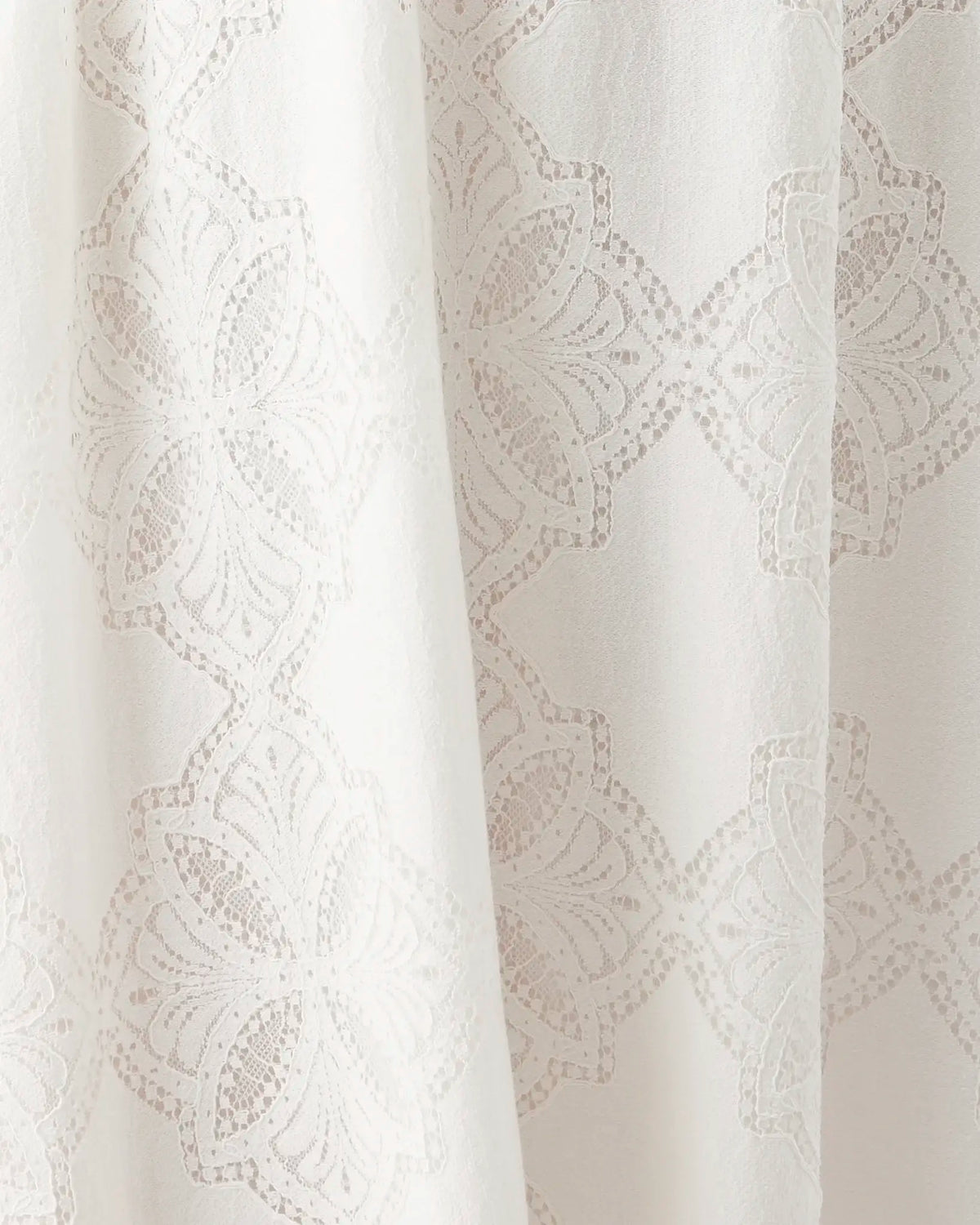 Close-up of a white or off-white fabric with a subtle tonal floral or lace-like woven pattern and soft vertical folds.