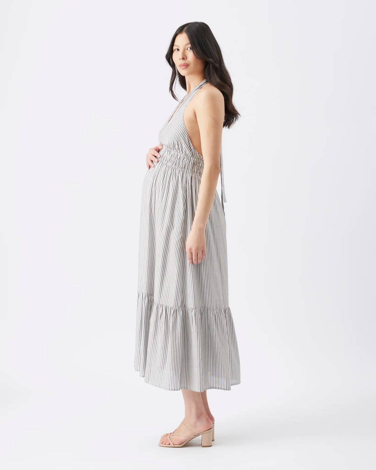 A pregnant woman wearing a light gray and white vertically striped halter midi dress with a smocked bodice and tiered skirt, standing in a studio setting.