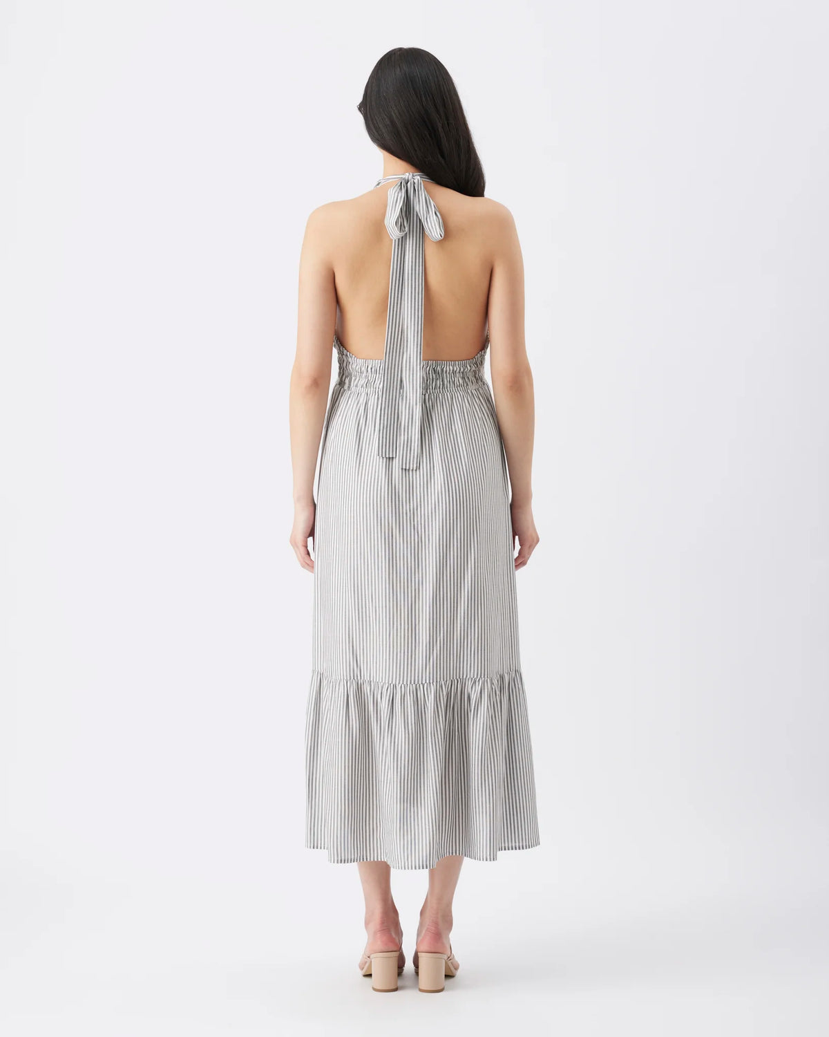A woman viewed from the back wearing a light gray and white vertically striped halter-neck midi dress with an open back and tie detail, paired with beige block-heel sandals, against a plain white studio background.