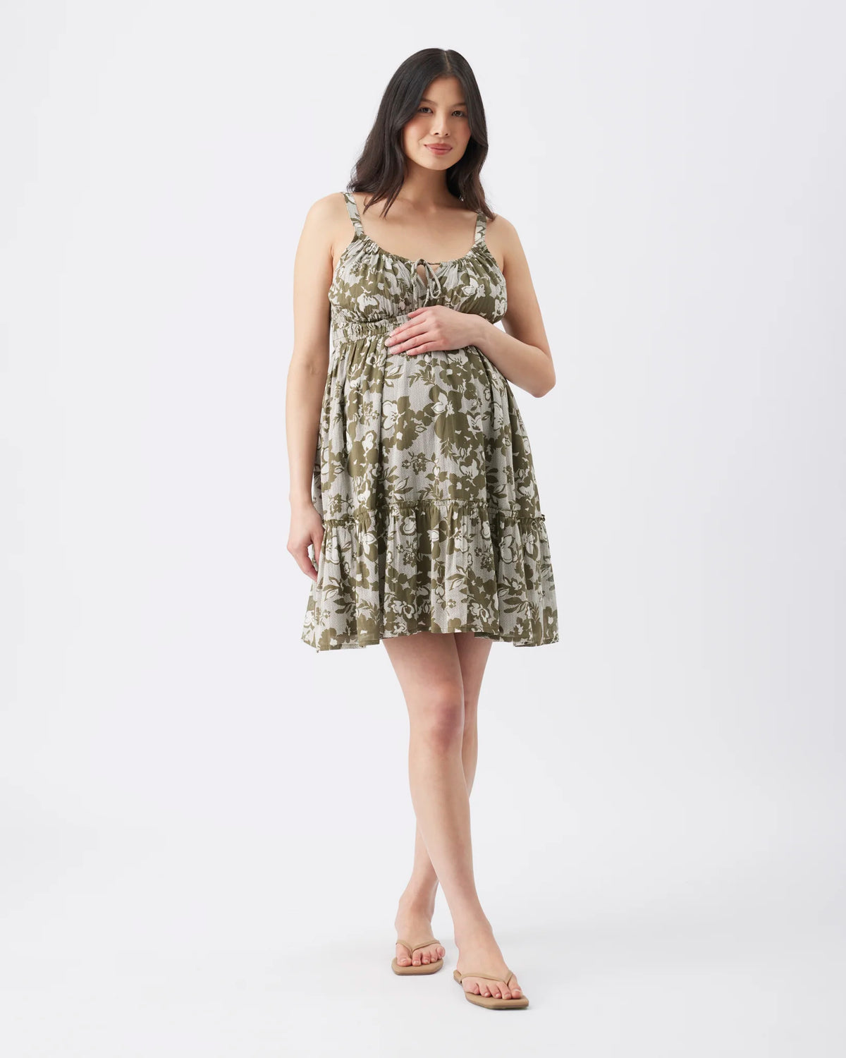 A woman modeling a sleeveless, tiered A-line maternity dress with a green and beige floral pattern, standing against a plain white studio background.