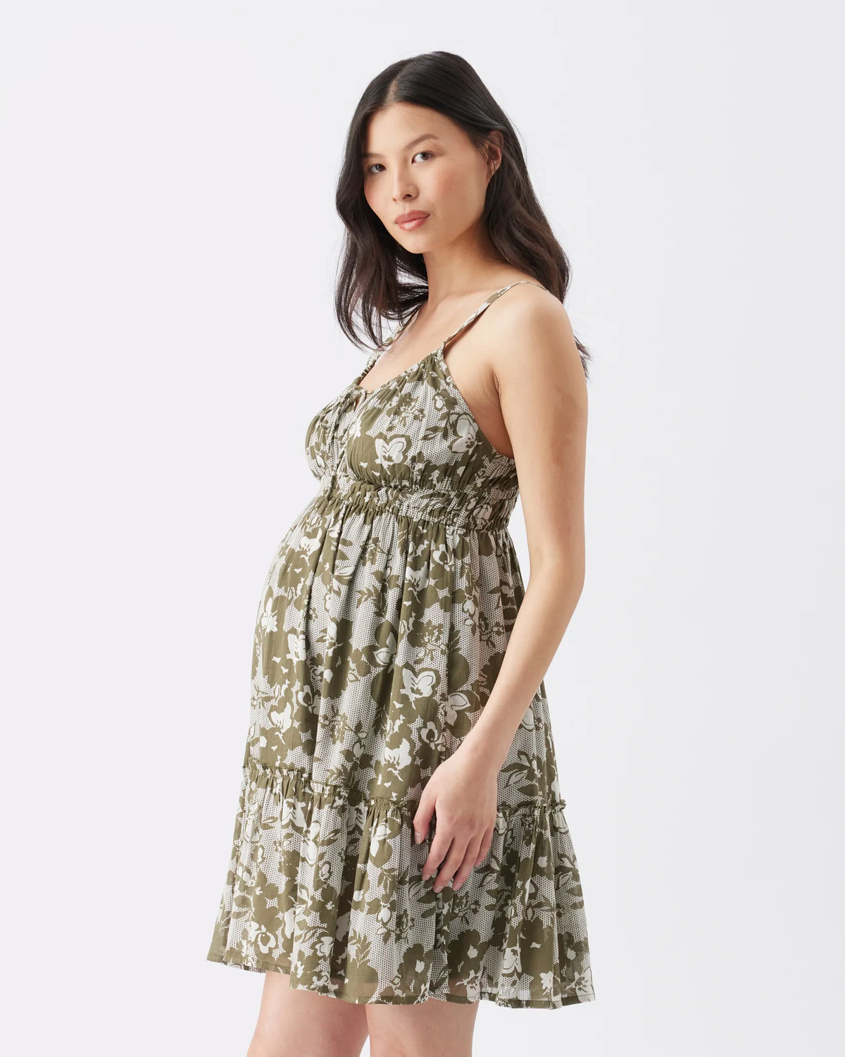 A pregnant model wearing a sleeveless, smocked babydoll maternity dress with an olive green and beige floral pattern, photographed against a solid white studio background.
