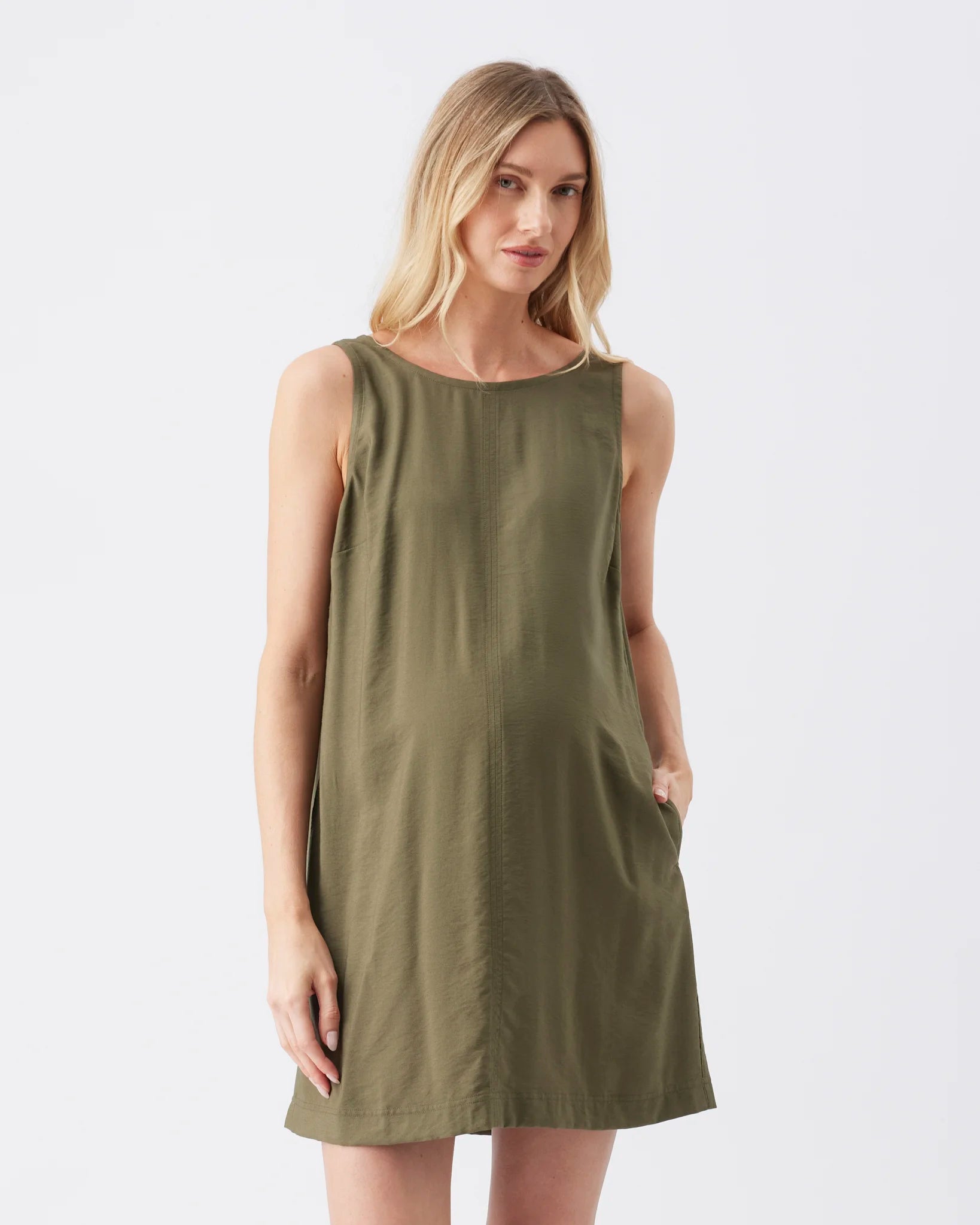 A woman modeling a sleeveless olive green shift dress against a plain light gray studio background.