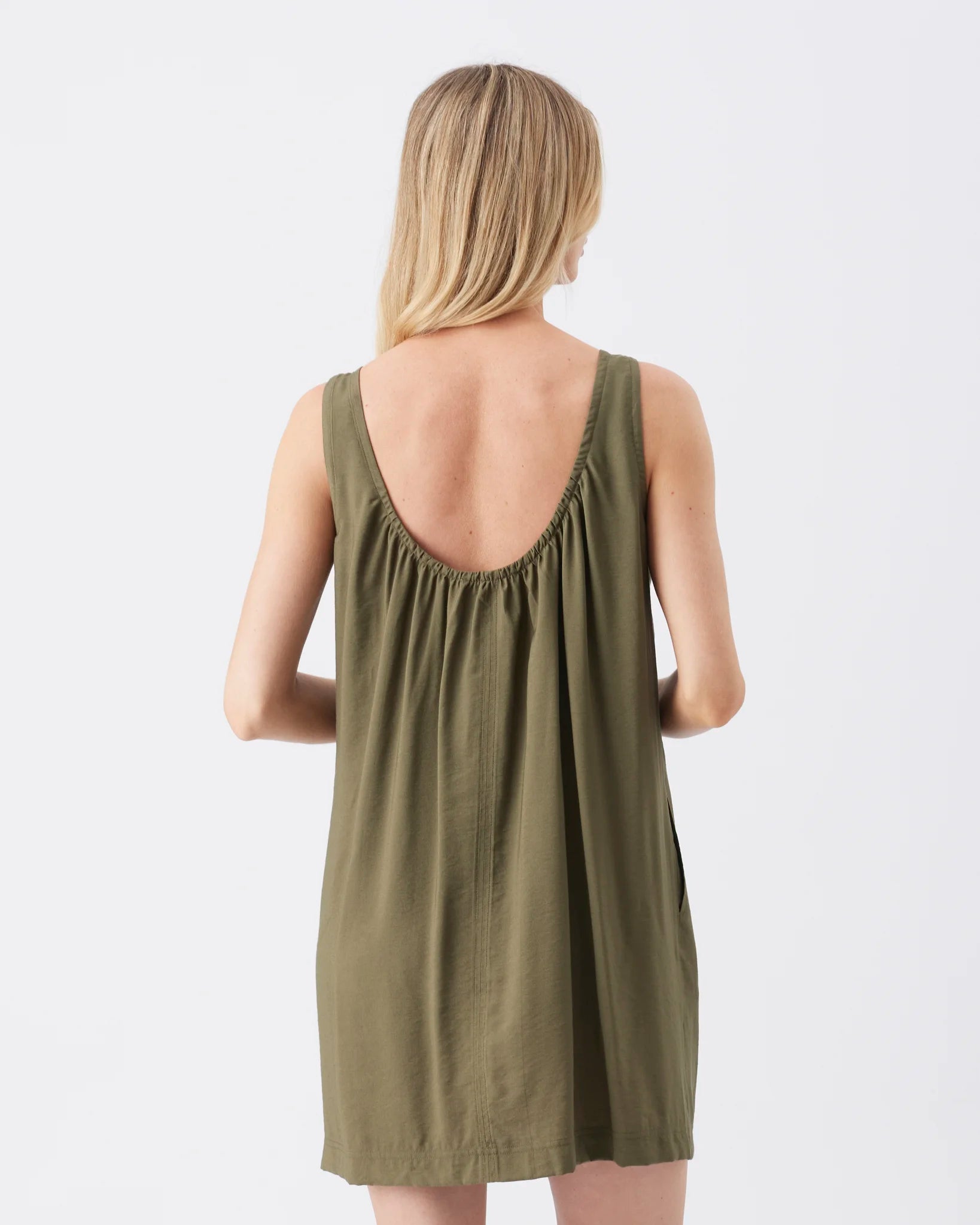 A woman viewed from the back wearing a sleeveless olive green dress with a gathered, low open back, set against a plain light gray studio background.
