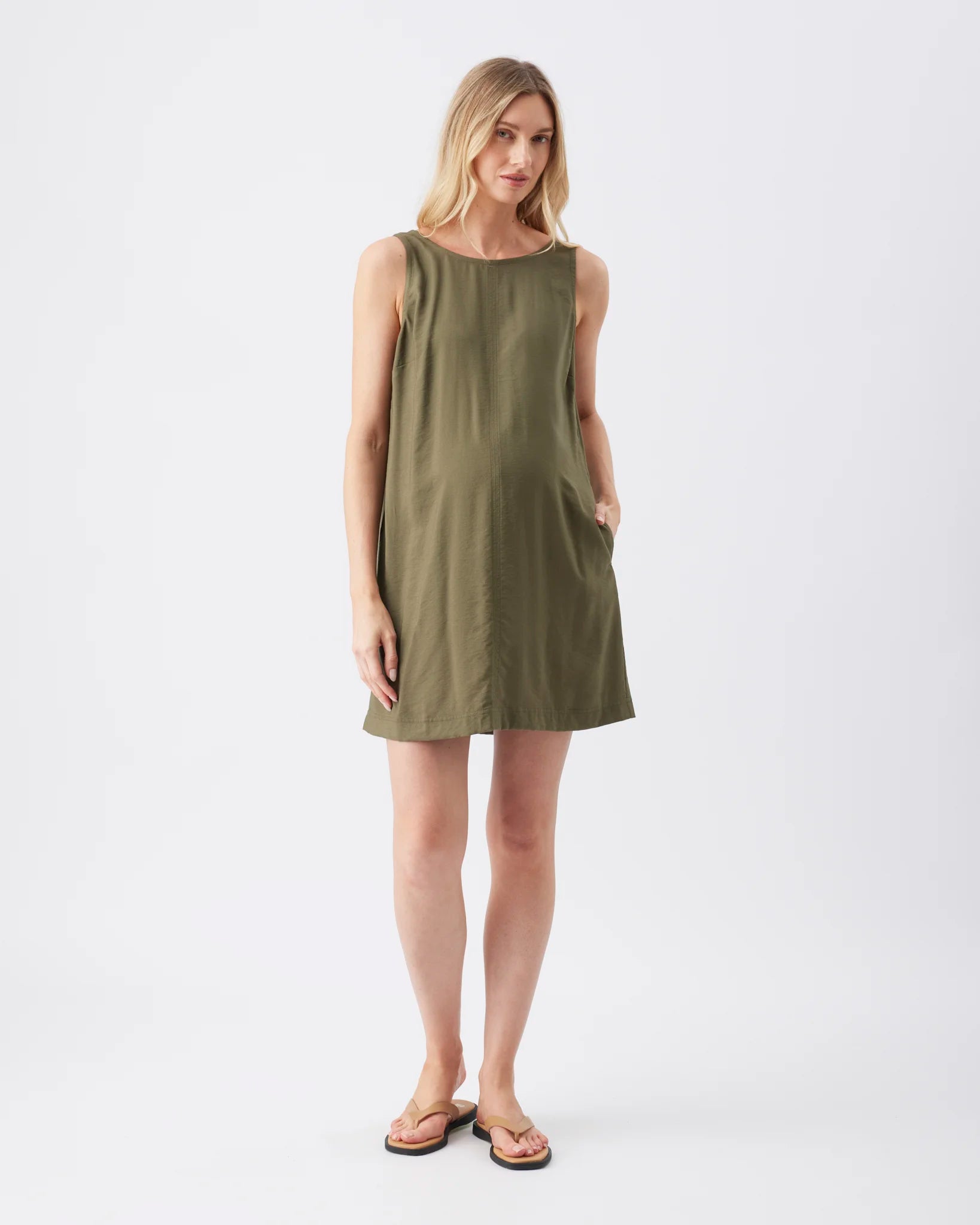 A woman modeling a sleeveless olive green shift dress with side pockets, paired with brown leather flip-flop sandals, against a plain white studio background.
