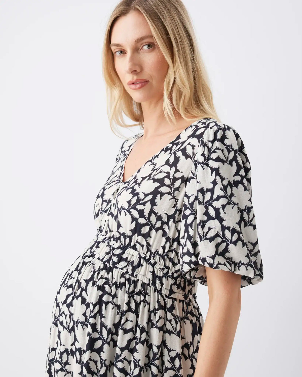 A pregnant model wearing a black and white floral print maternity dress with puff sleeves and a gathered waist, photographed against a clean white studio background.