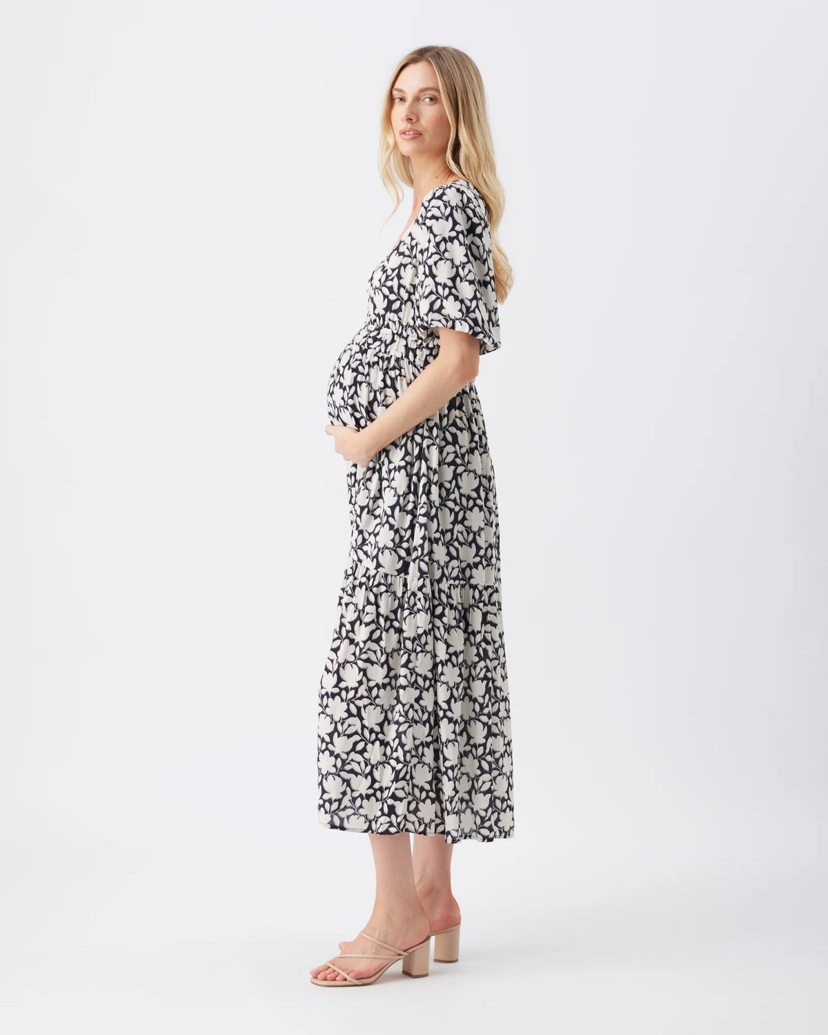 A pregnant woman wearing a black and white floral print midi dress with flutter sleeves, standing in profile against a plain white studio background.