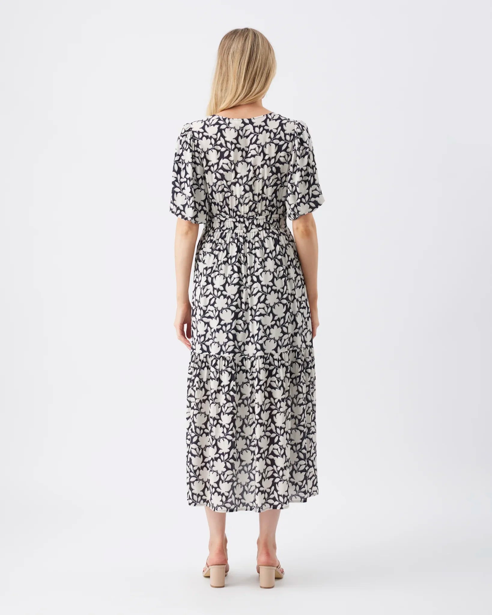 A woman viewed from the back wearing a short-sleeve midi dress with a black and white floral pattern, paired with beige block-heel sandals, against a plain white studio background.