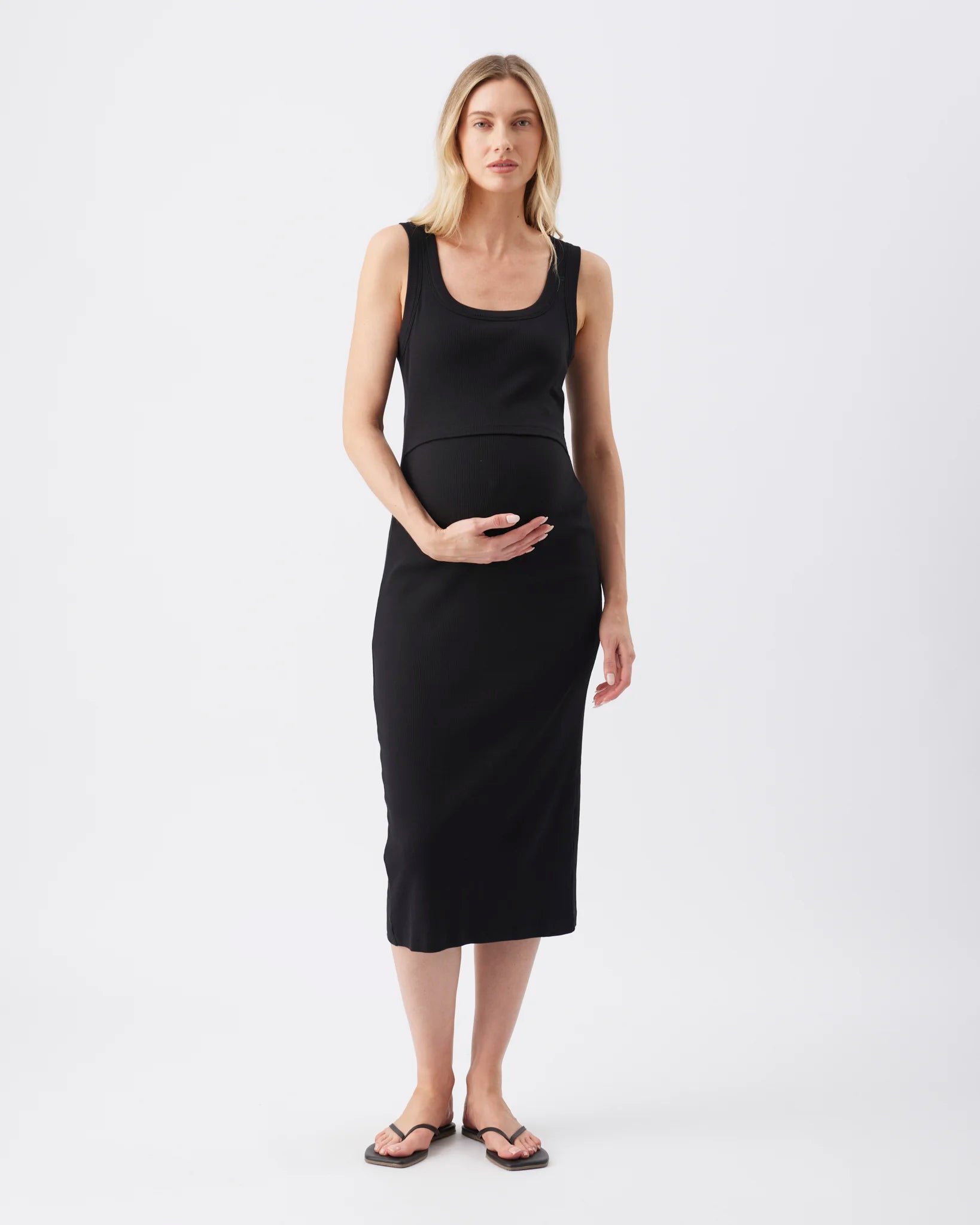 A pregnant woman modeling a black sleeveless maternity dress with a scoop neckline and a fitted silhouette, standing against a plain white studio background.