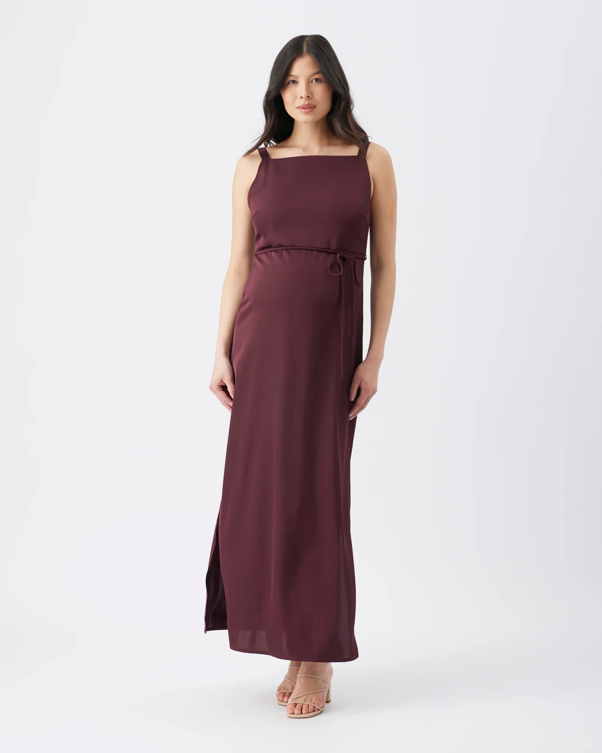 A woman modeling a sleeveless, floor-length mauve-burgundy dress with a square neckline, thin shoulder straps, a self-tie waist belt, and side slits, photographed against a plain light gray studio background.