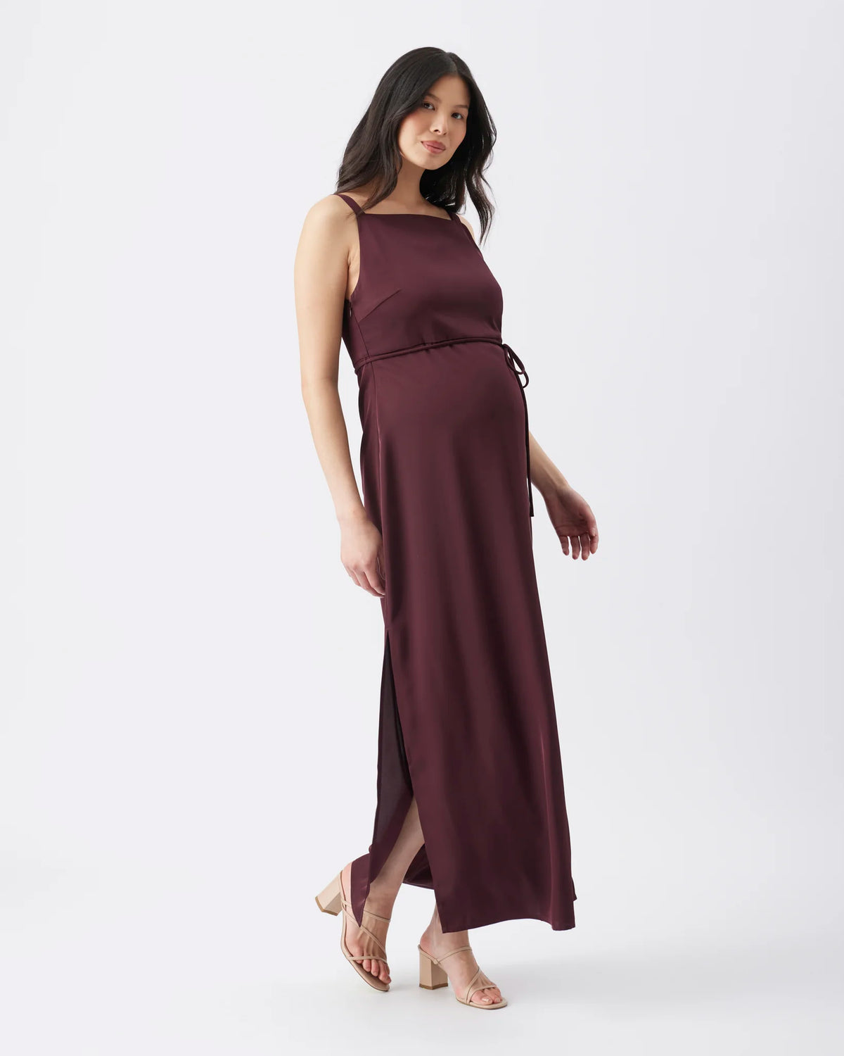 A pregnant woman modeling a sleeveless, dark plum-burgundy maxi maternity dress with a side slit and a matching tie belt at the waist, set against a plain white studio background.
