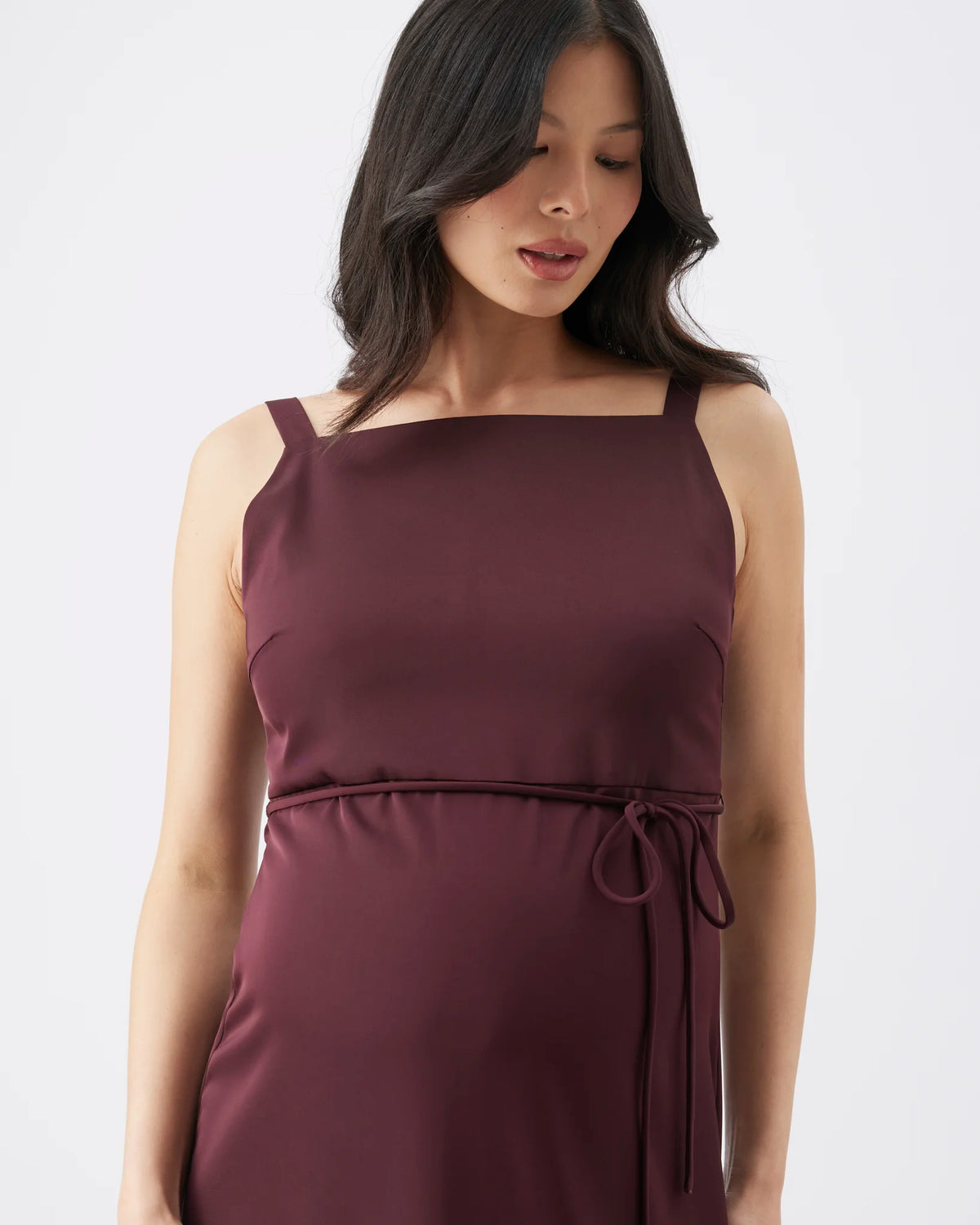 A woman wearing a sleeveless, scoop-neck maternity dress in a deep plum purple color, featuring thin shoulder straps and a drawstring waist, set against a plain light gray background.