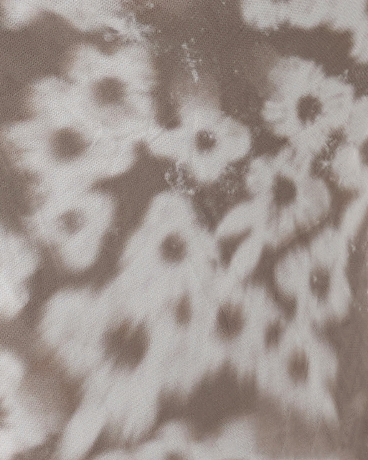 A close-up of a tie-dye fabric pattern featuring organic, flower-like shapes in shades of taupe brown and off-white.