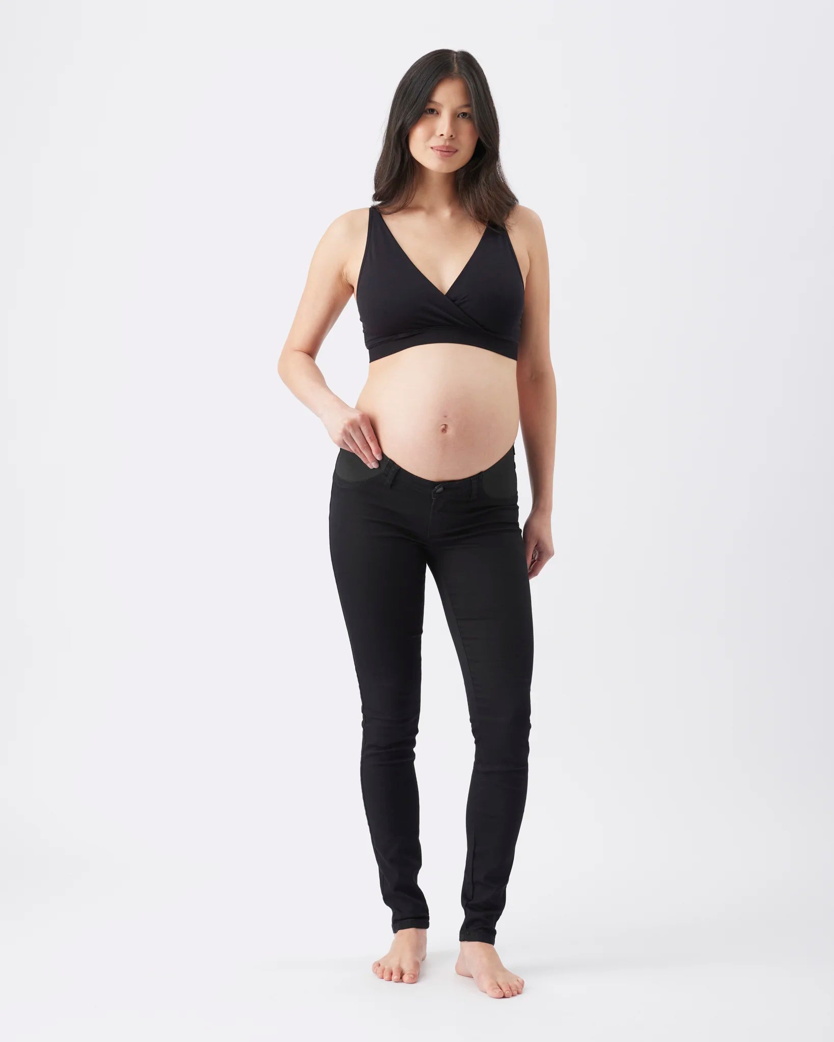 A pregnant woman stands barefoot against a plain white studio background, wearing a black maternity wrap-style bra top and black maternity jeans.