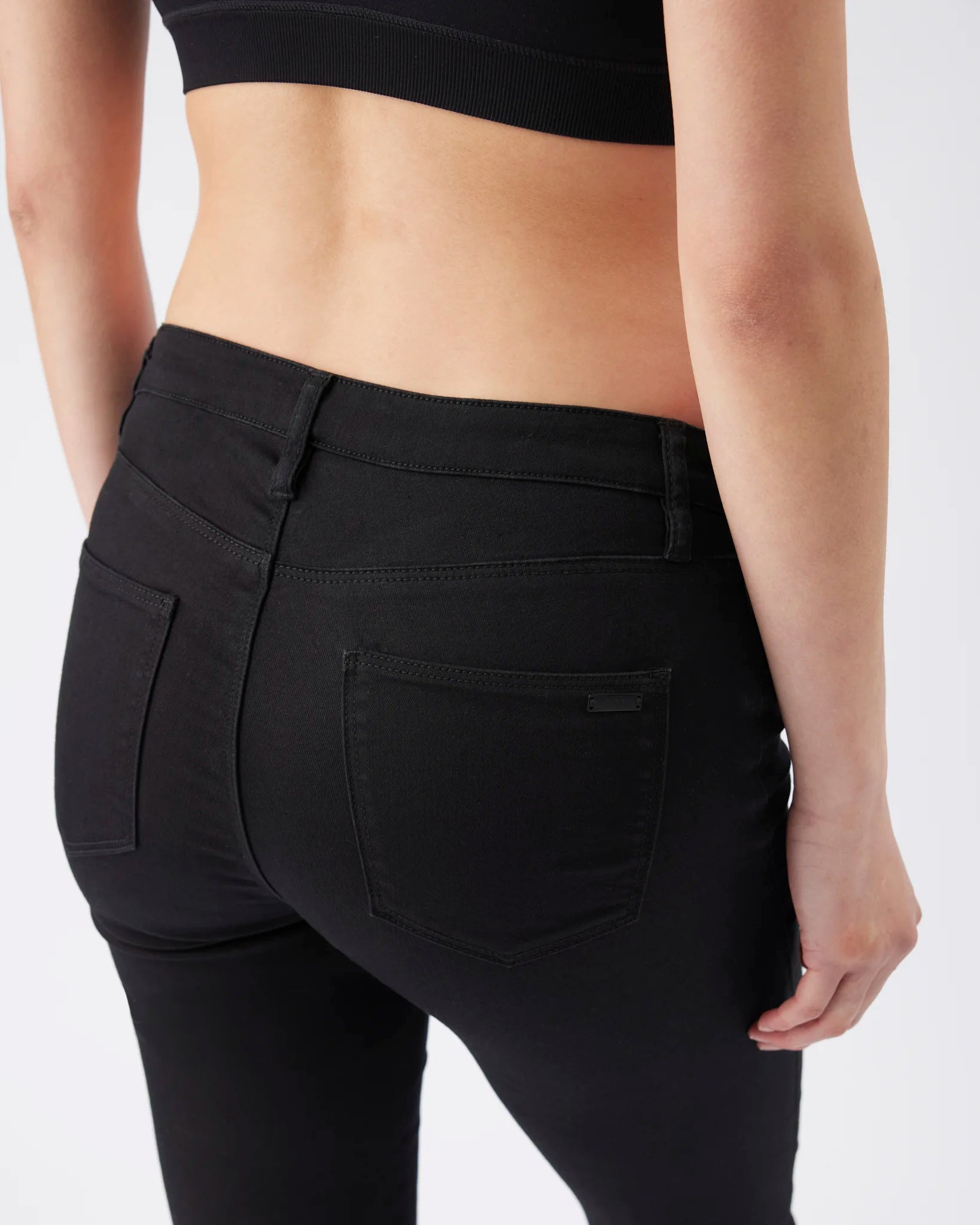 Rear view of a model wearing black high-waisted skinny jeans with a visible waistband and back pocket details, paired with a black crop top against a light gray studio background.