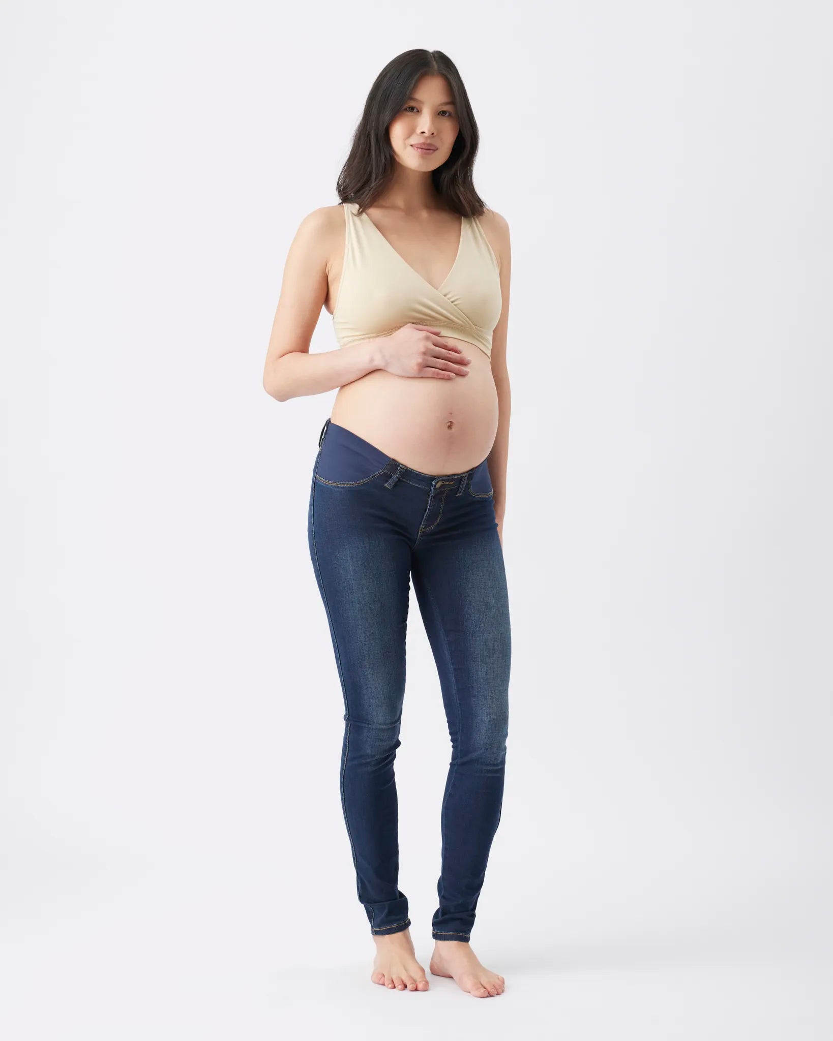 A pregnant woman stands barefoot against a plain white studio background, wearing a light beige wrap-style halter top and dark blue skinny jeans.