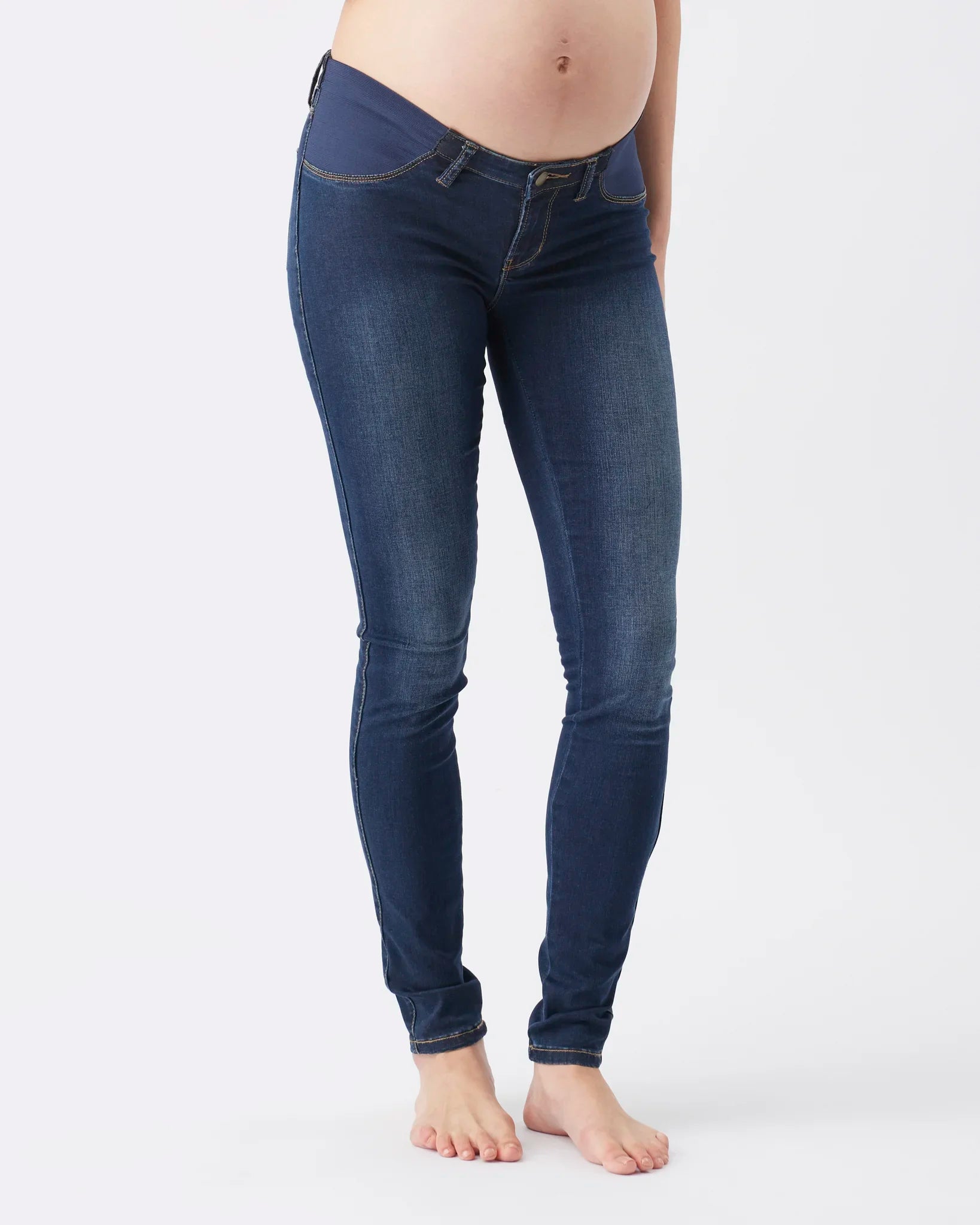 A pregnant model wearing dark blue maternity skinny jeans, shown from the waist down against a plain white studio background.