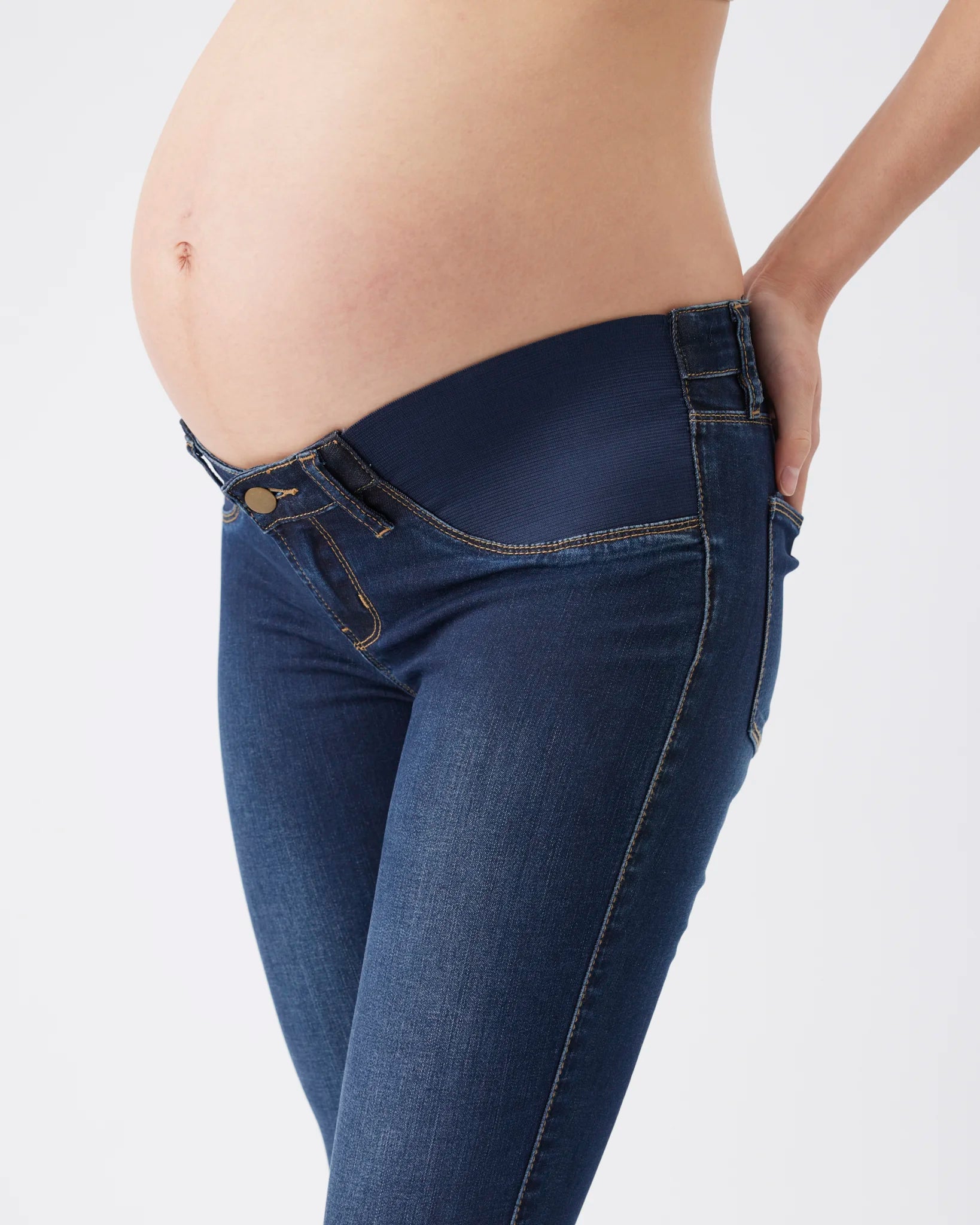 A close-up of a pregnant model wearing dark blue maternity jeans with a wide, comfortable waistband, shown from the side against a plain white background.