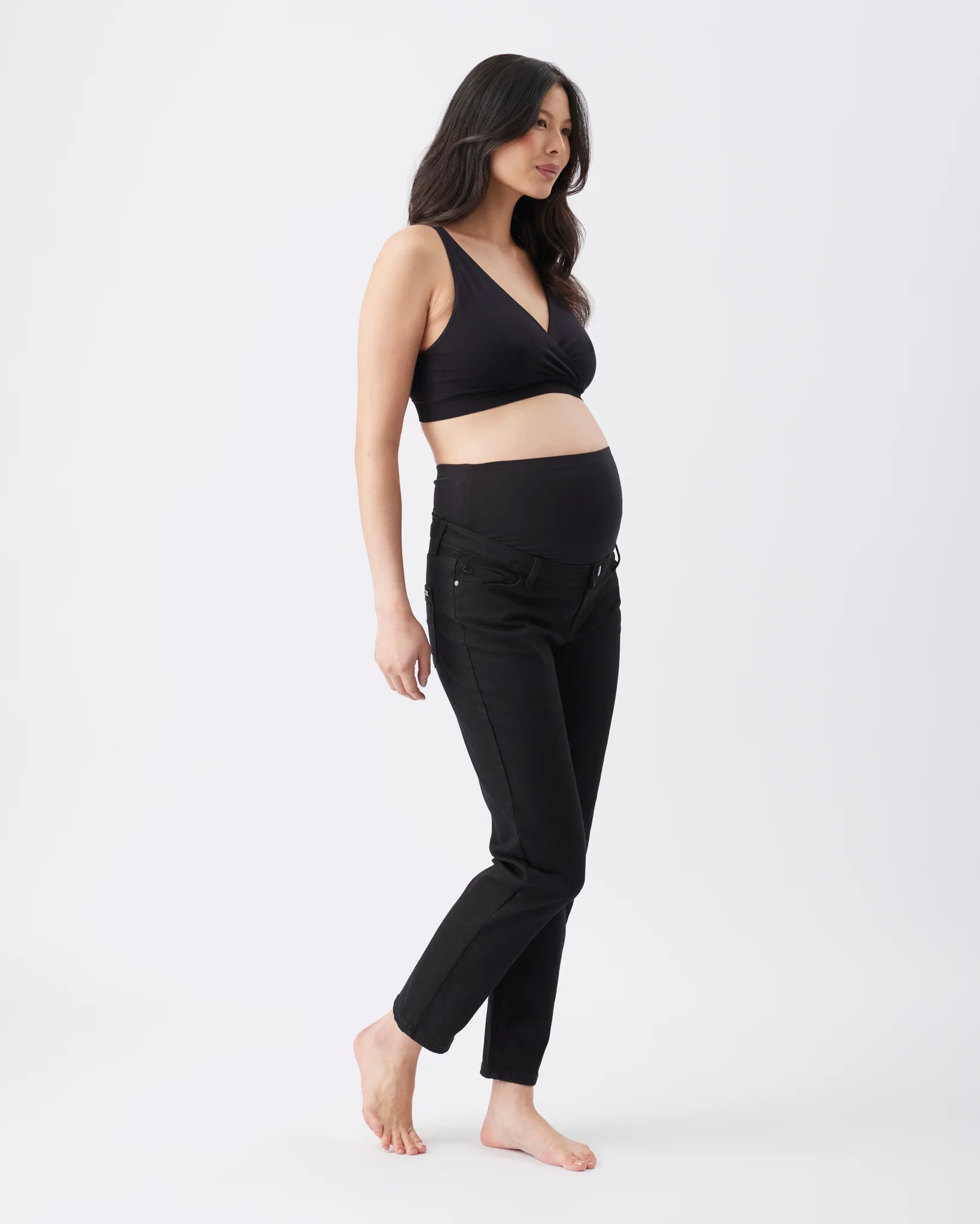 A pregnant woman stands in profile wearing a black wrap-style maternity bra and black maternity jeans against a plain white studio background.