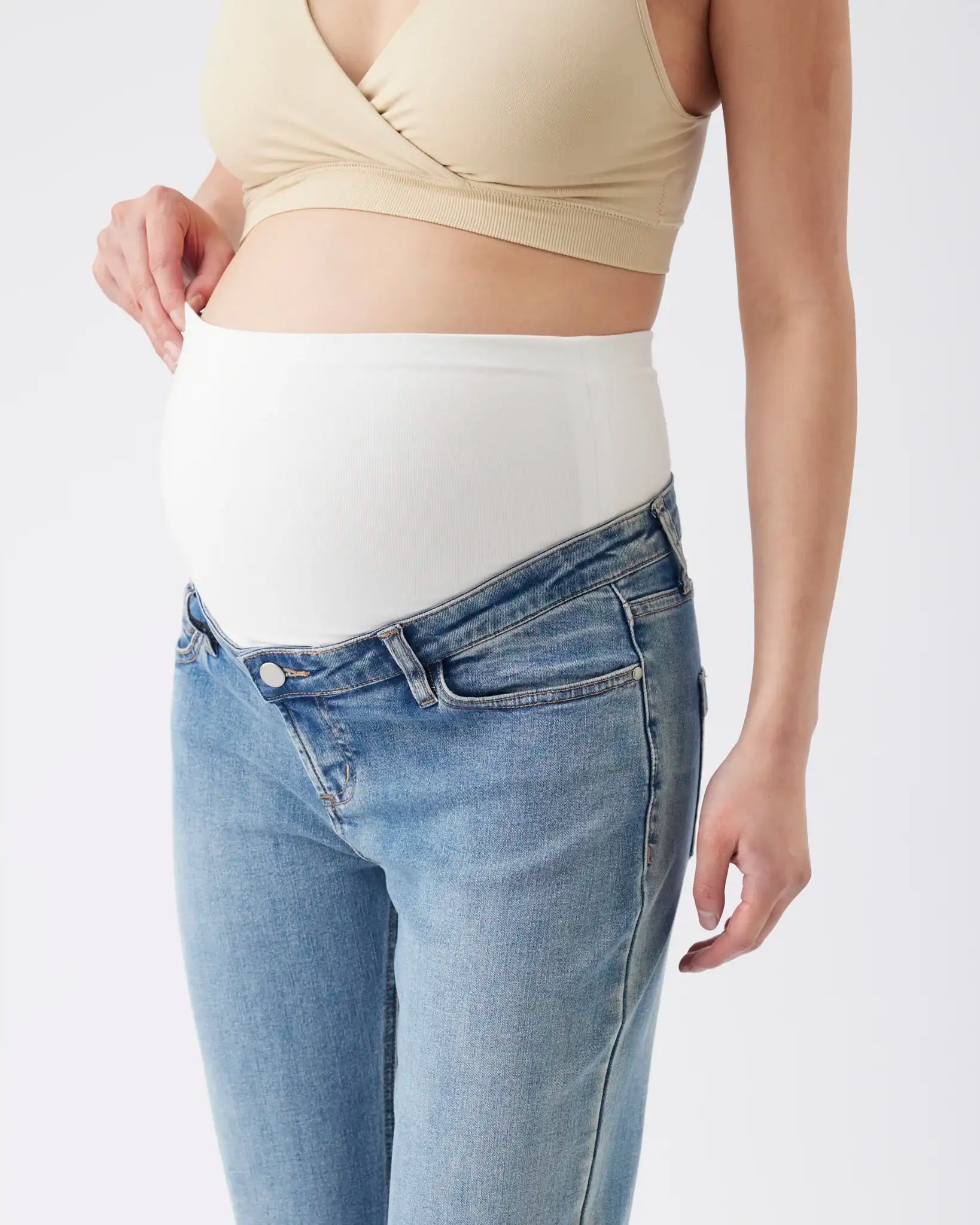 A pregnant model wearing a white maternity belly band under a beige wrap-style top and light blue jeans, shown from the chest down against a plain light gray background.