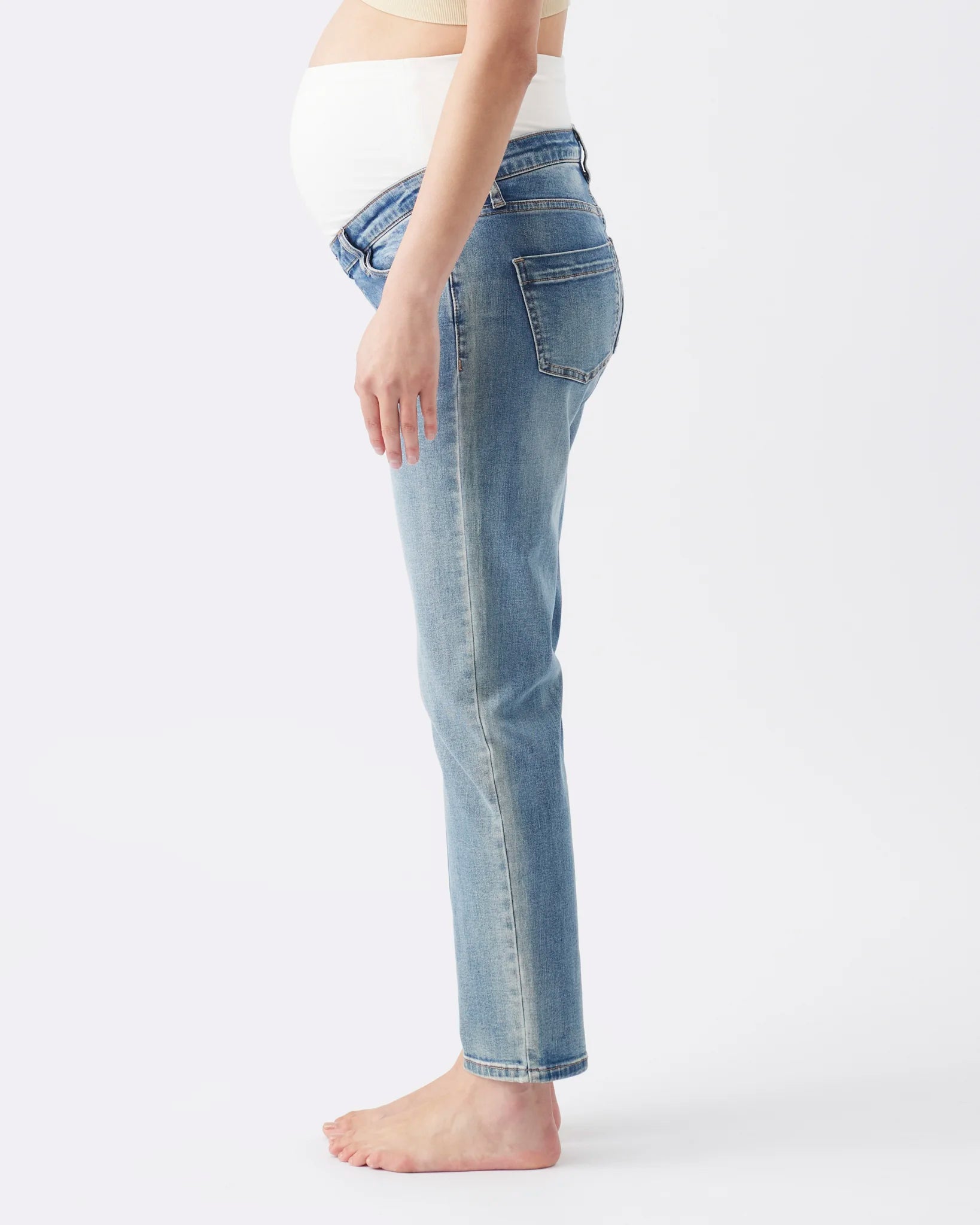 Side view of a pregnant model wearing light-wash blue maternity jeans and a white maternity top, standing barefoot against a plain white studio background.