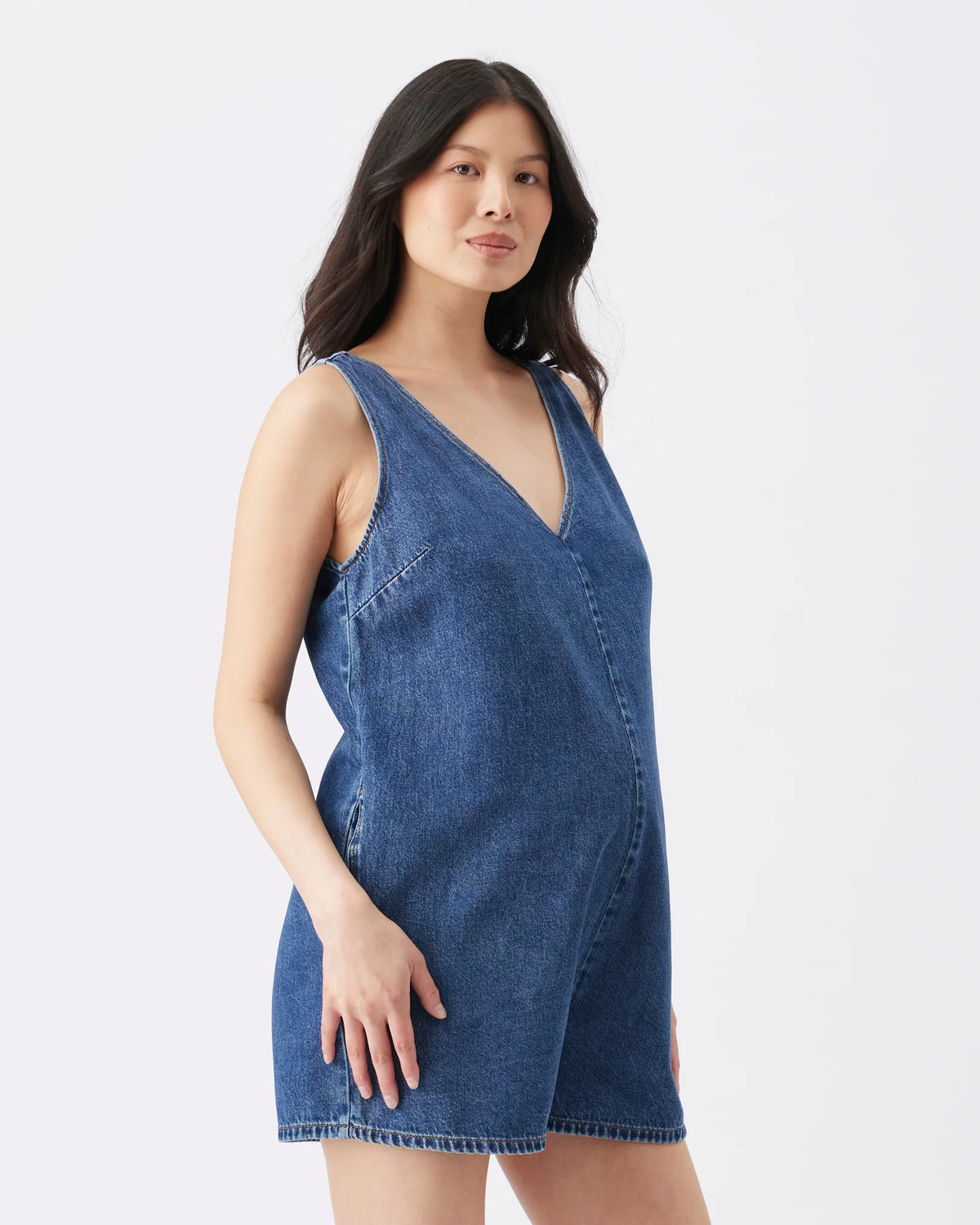 A woman modeling a sleeveless blue denim romper with a V-neckline, photographed against a clean light gray studio background.