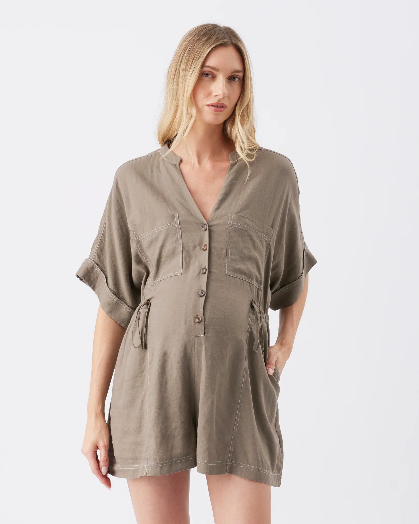 A woman modeling a short-sleeve, button-front romper in a muted taupe-brown color against a plain light gray studio background.