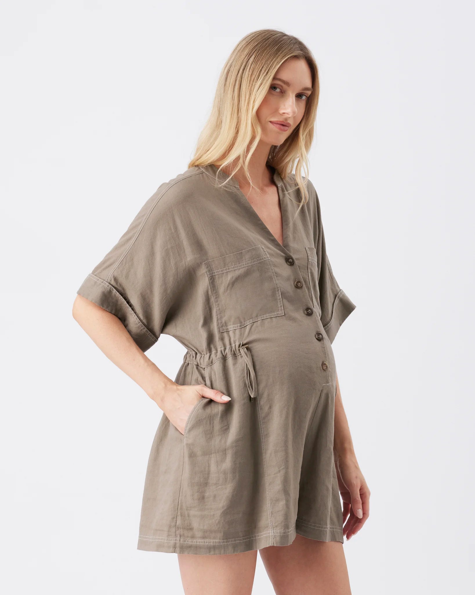 A blonde model wearing a short-sleeved, button-front linen romper in a muted taupe-brown color, photographed against a clean white studio background.