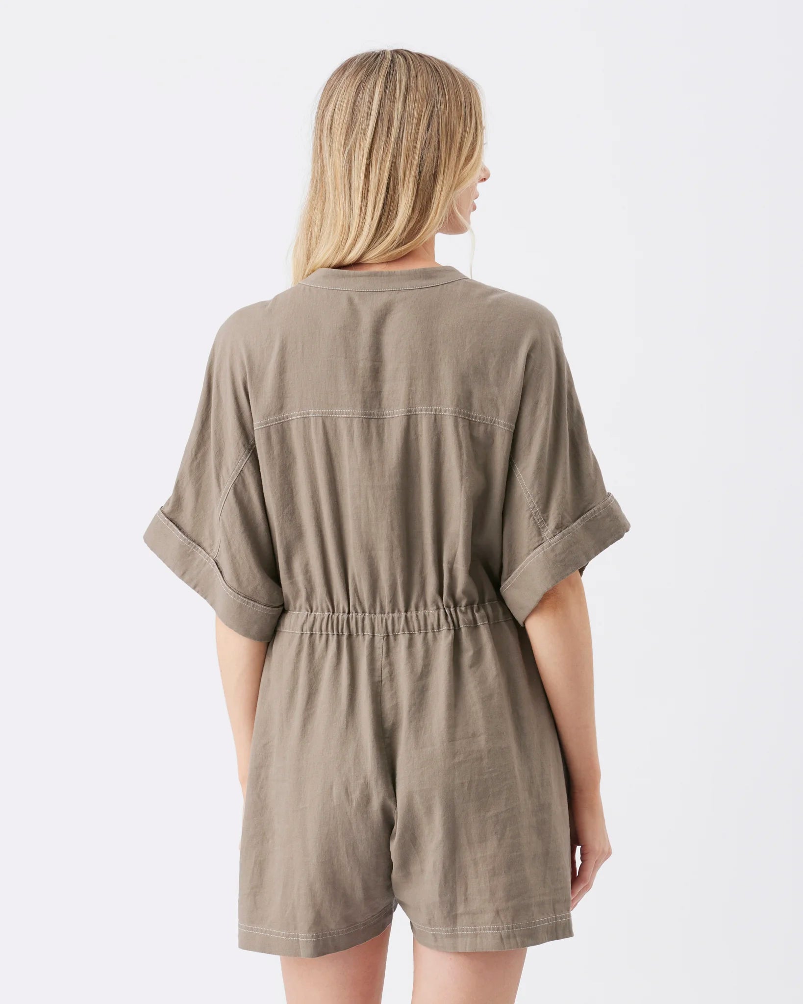 A woman viewed from the back wearing a loose taupe-brown short romper with rolled sleeves and an elastic waist, against a plain light gray studio background.
