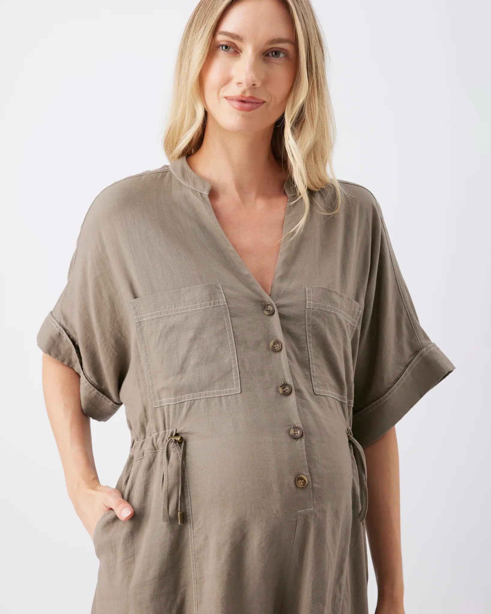 A woman wearing a short-sleeve, button-front linen-like dress or tunic in a muted taupe-brown color, photographed against a clean light gray studio background.