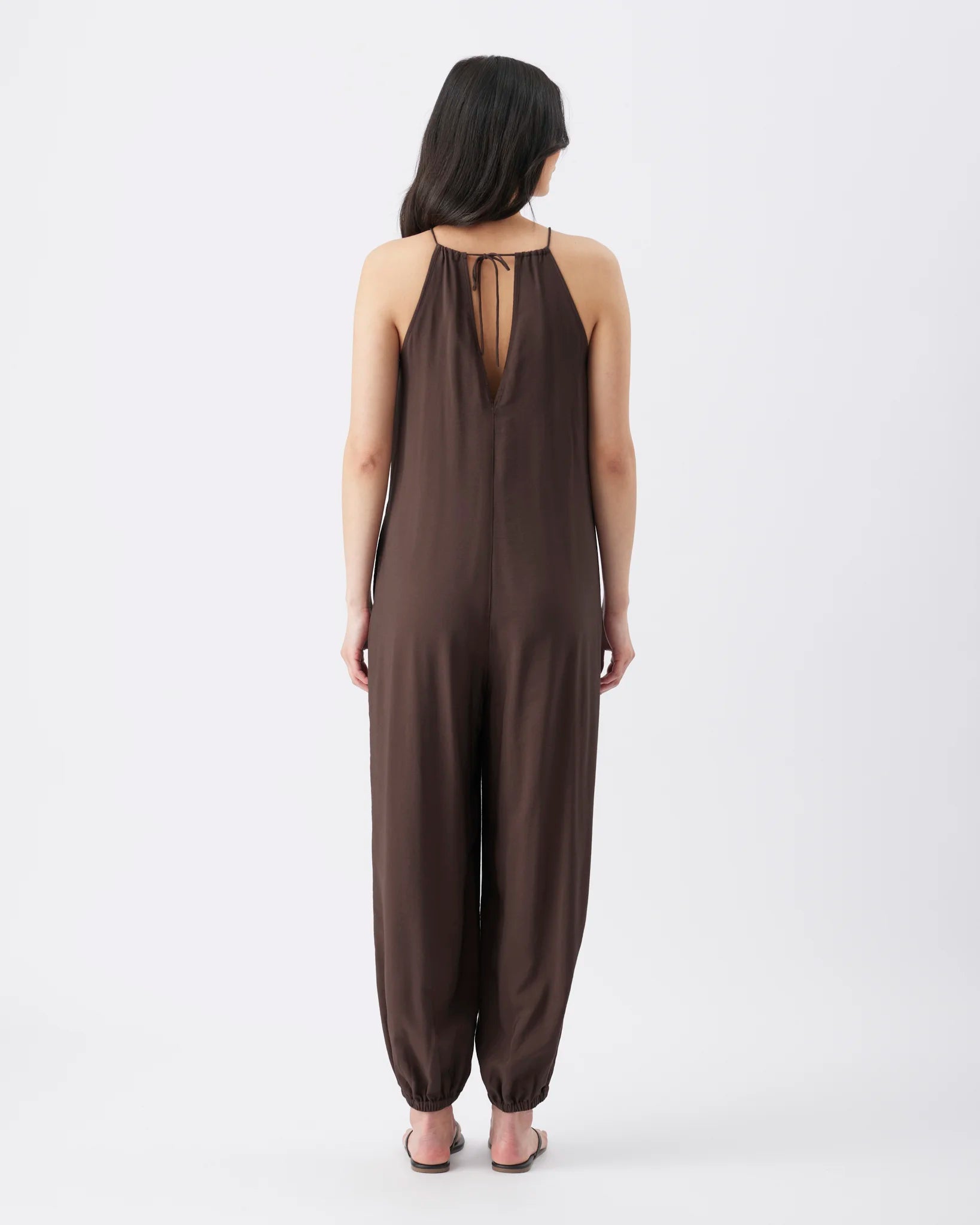 A woman viewed from the back wearing a dark brown sleeveless jumpsuit with a tie-back neckline and loose, wide-leg pants, standing against a plain light gray studio background.