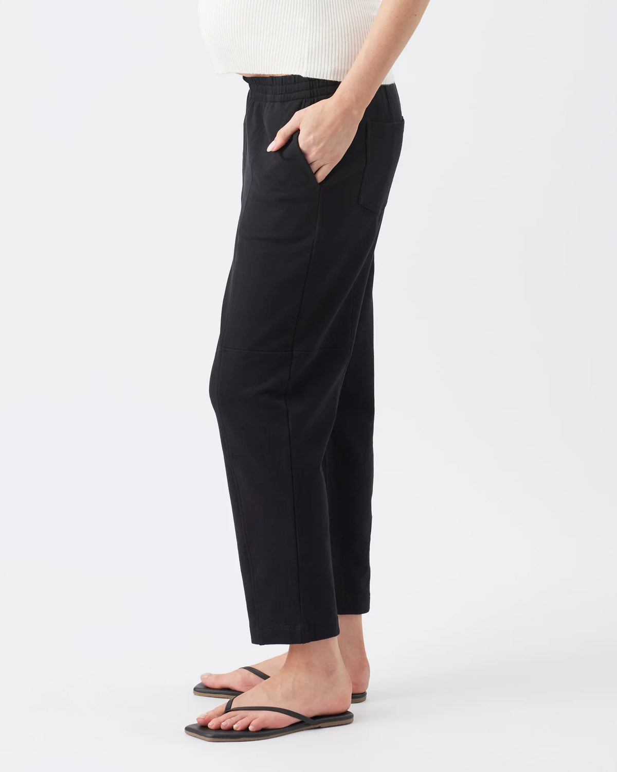 A person wearing black cropped pants with side pockets, paired with a white ribbed top and black flip-flops, shown against a plain light gray studio background.