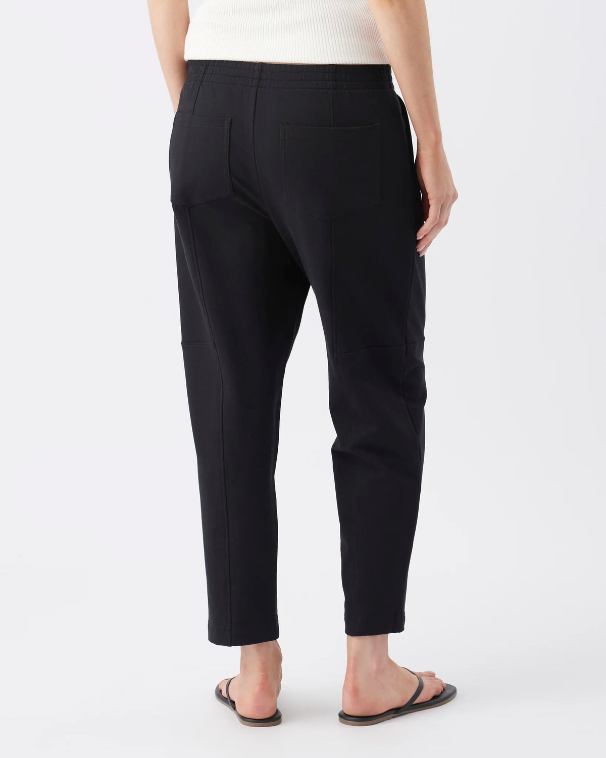 Rear view of a person wearing black cropped pants with a white ribbed top and dark sandals, photographed against a clean light gray studio background.