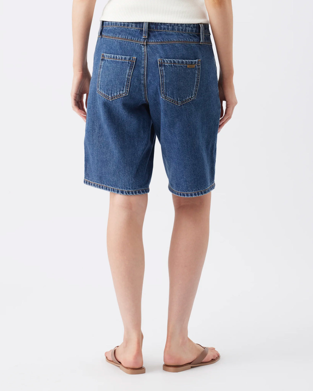 A person shown from the back wearing blue denim shorts with a cream ribbed top and brown sandals, photographed against a clean light gray studio background.