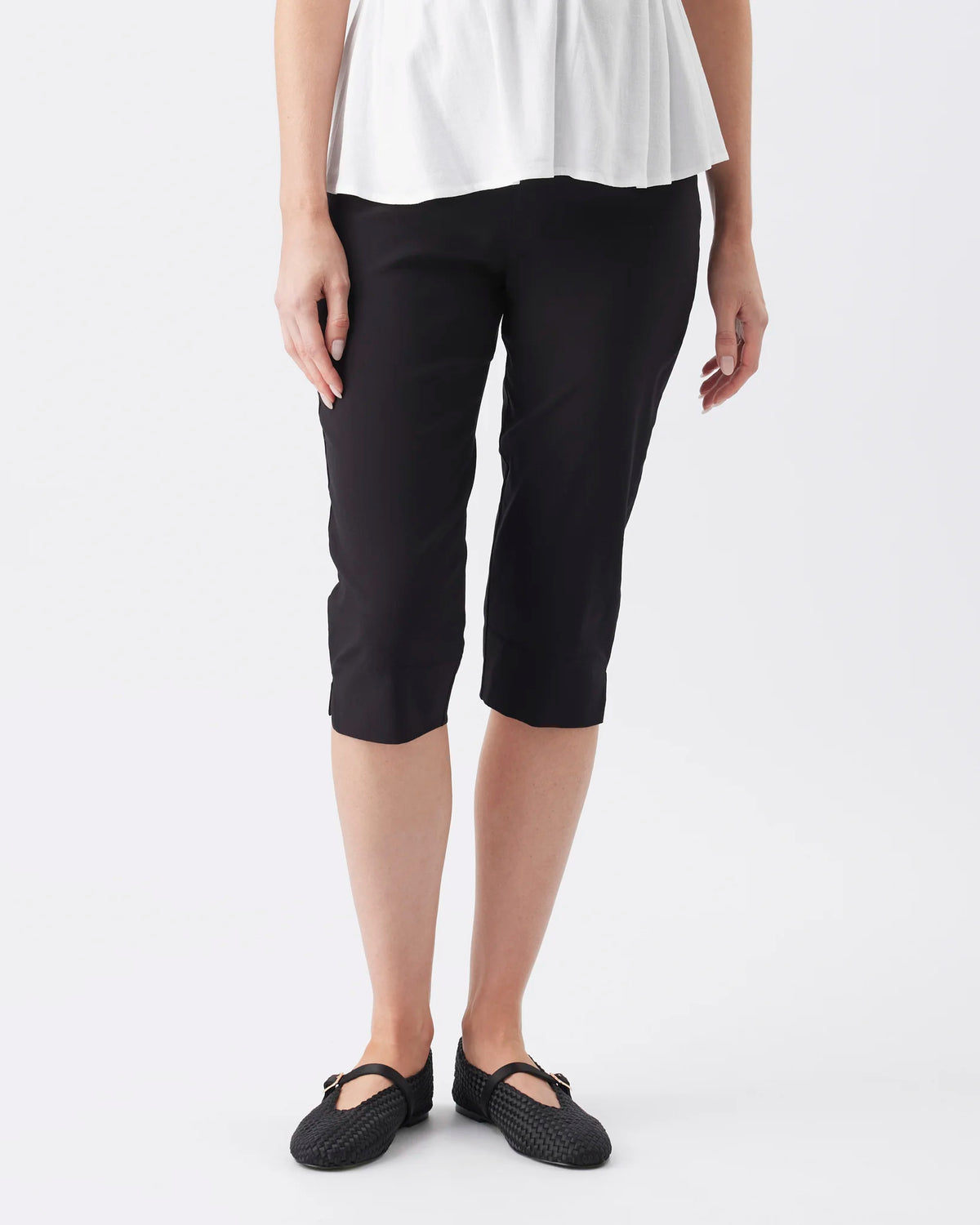 A model wearing black cropped capri pants with a white top and black flat shoes, shown from the waist down against a light gray studio background.