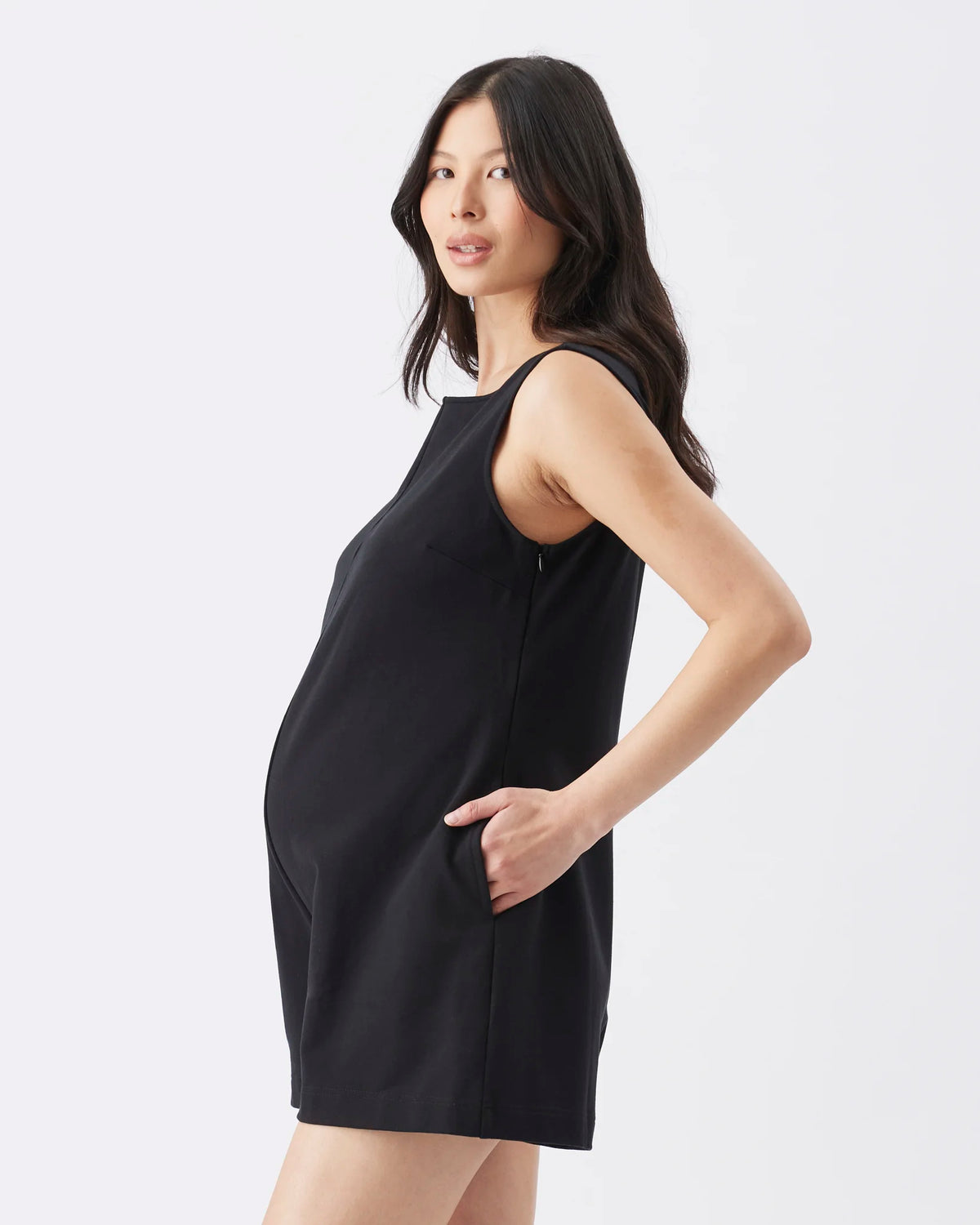 A woman modeling a sleeveless black maternity dress with side pockets, shown from a three-quarter back angle against a light gray studio background.
