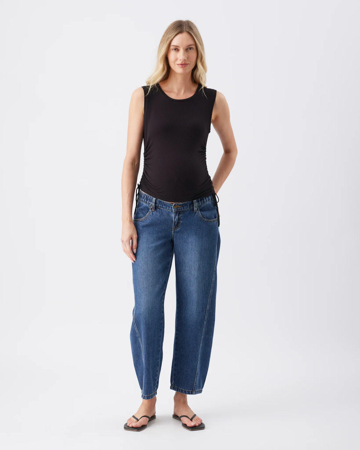 A woman modeling a black sleeveless top and blue wide-leg cropped jeans with dark sandals, standing against a plain light gray studio background.