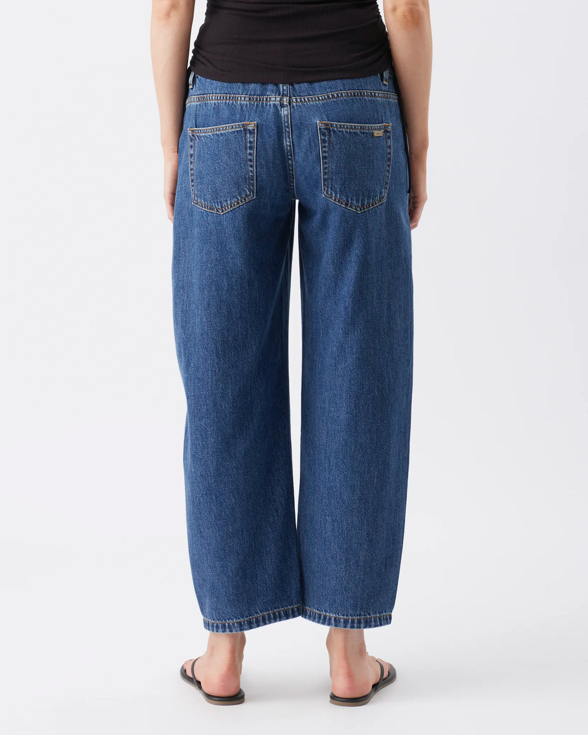 Rear view of a person wearing wide-leg cropped blue denim jeans with a black top and dark sandals, photographed against a light gray studio background.