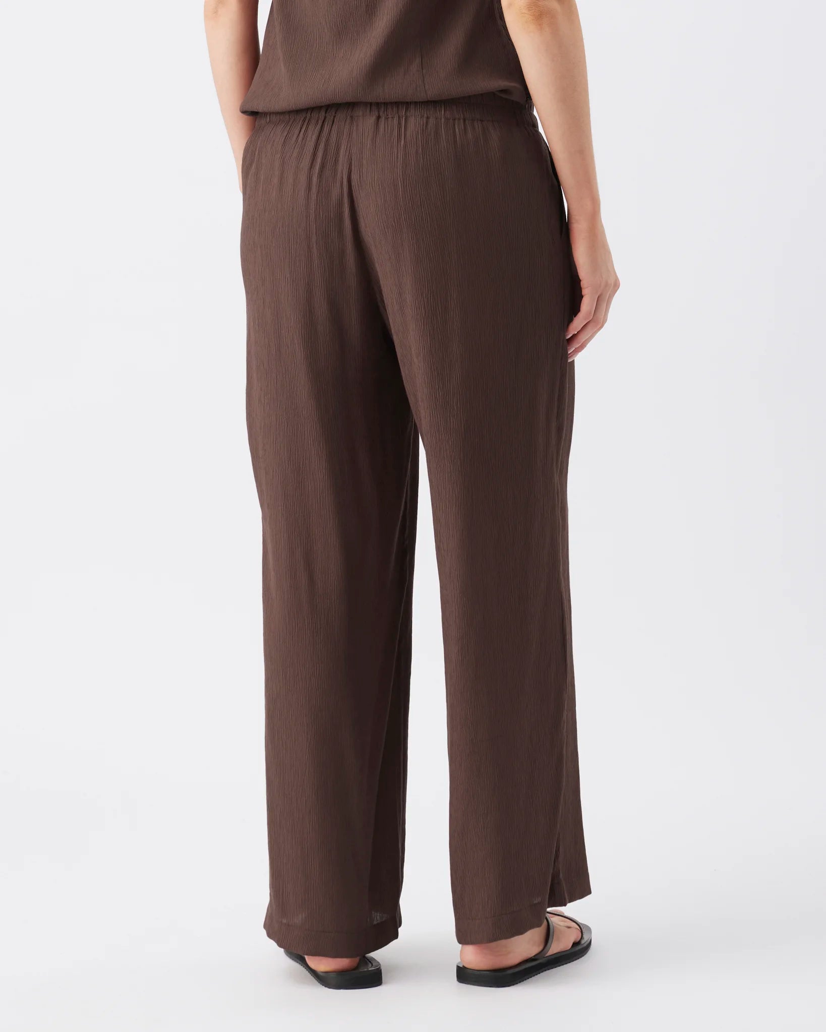 A person shown from the back wearing loose, wide-leg brown pants and black sandals against a clean white studio background.