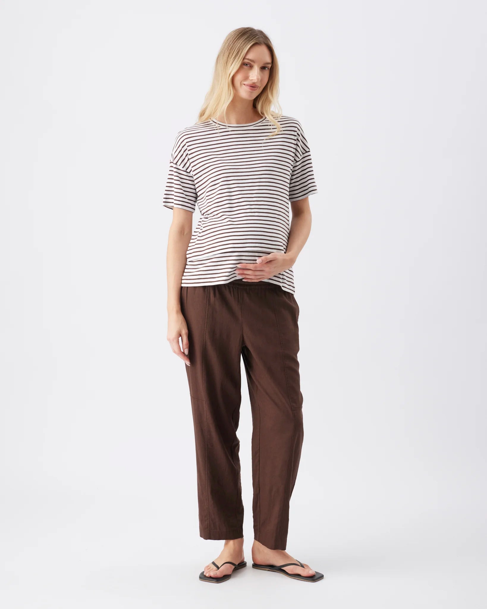 A woman modeling a short-sleeve striped t-shirt and dark brown wide-leg pants against a plain light gray studio background.