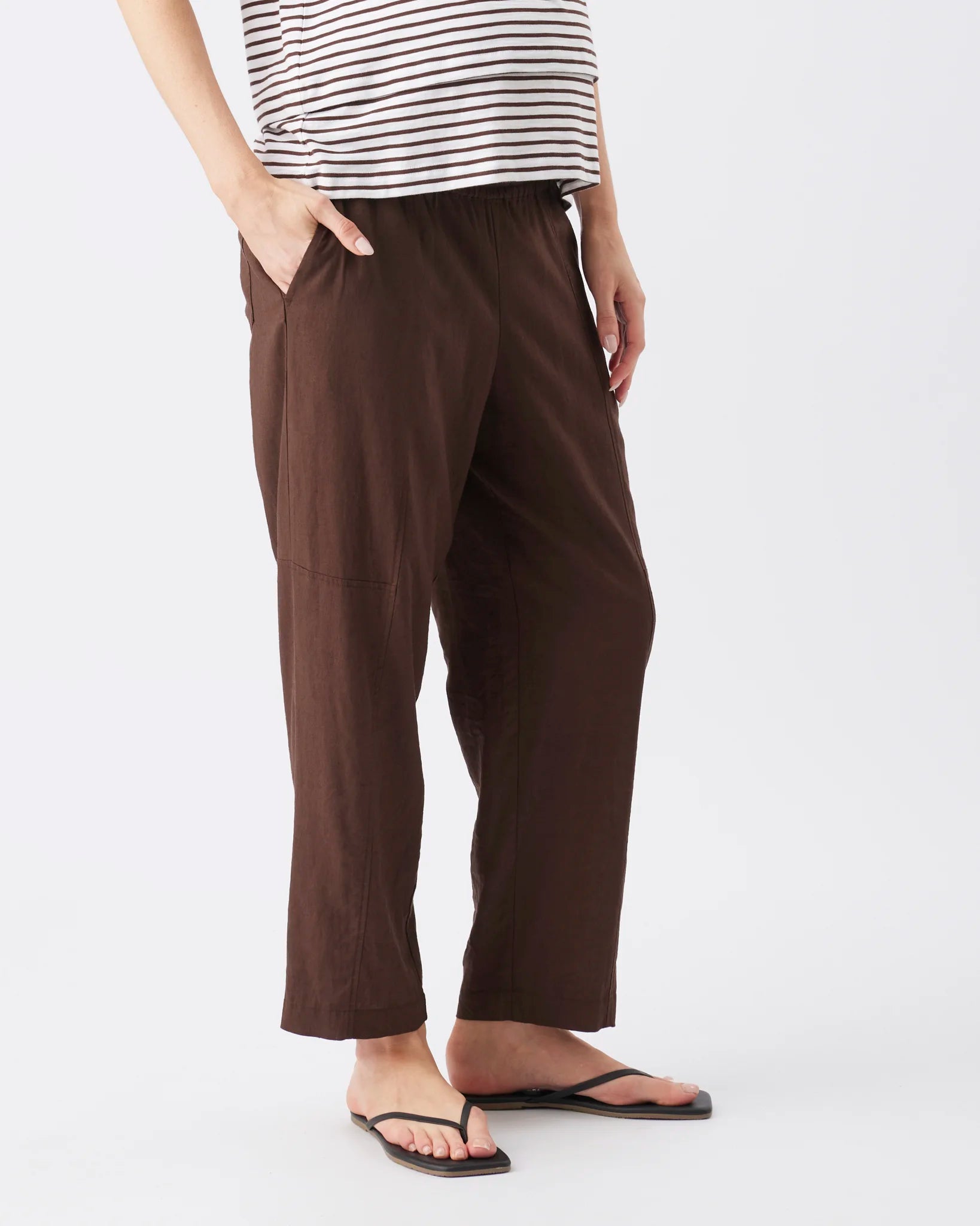 A person wearing dark brown cropped pants with side pockets, paired with a black and white striped top and black flip-flop sandals, shown against a plain light gray studio background.